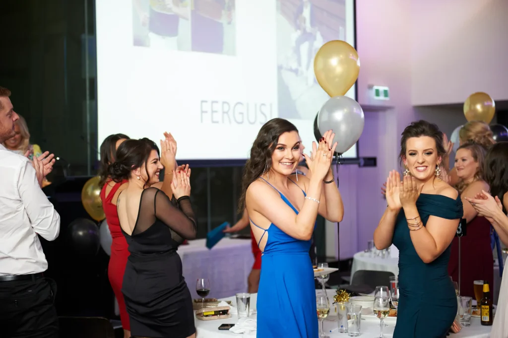 svg%3E Guests share a moment of joyful applause at the Padraig Pearses GAC Awards Night & Gala Dinner at the MCG. Women in elegant evening gowns, including a bright blue dress and a dark teal off-shoulder dress, clap and smile warmly as they engage with the event. A man in a white shirt and waistcoat joins in, while dinner tables, gold and silver balloons, and a projection screen create a festive, formal atmosphere celebrating awards and meaningful connections.