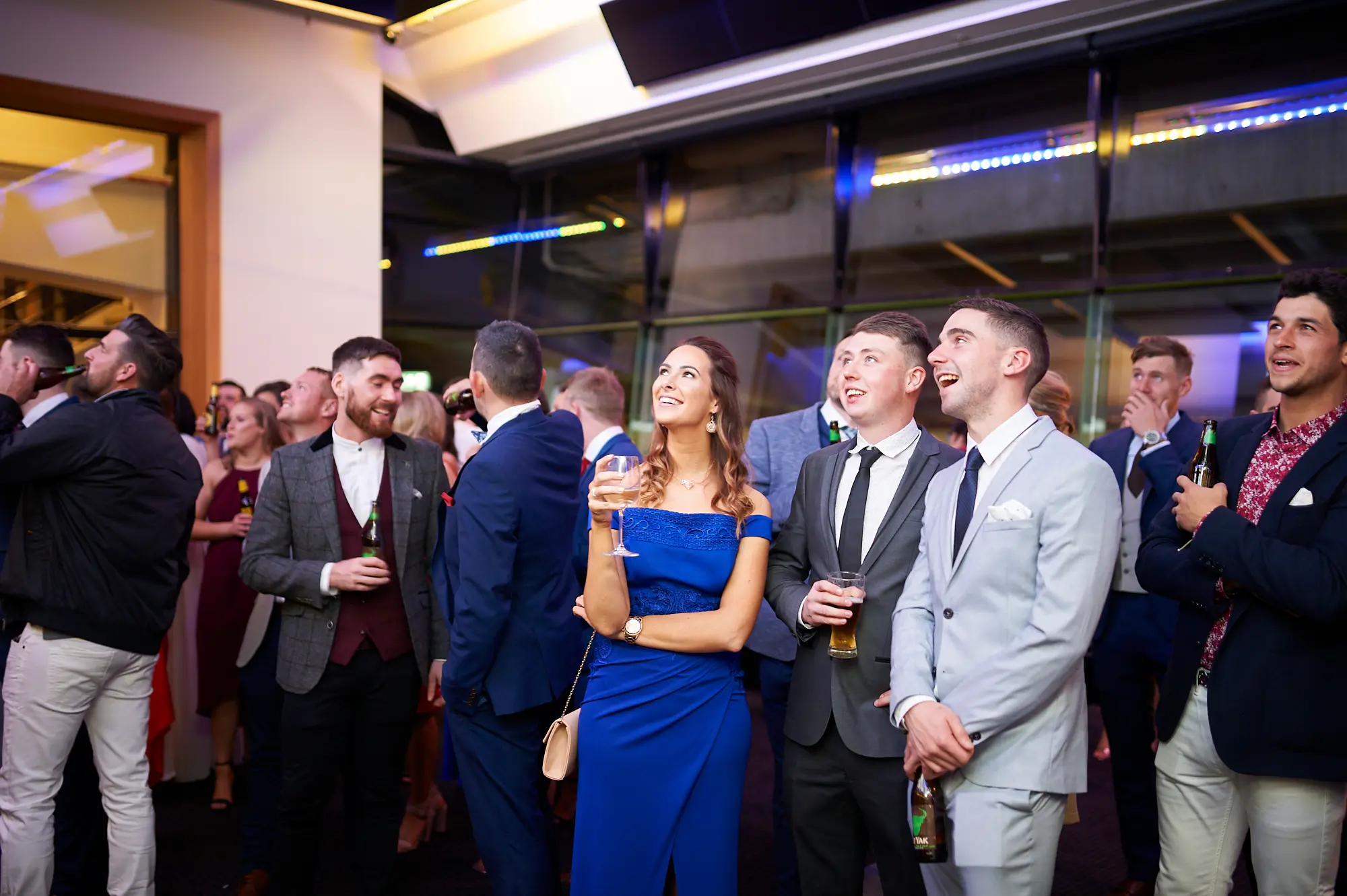 padraig pearses gac awards night mcg guests arrival blue dress engaged A group of guests watches attentively during the arrival period of the Padraig Pearses GAC Awards Night & Gala Dinner at the MCG. A woman in a vibrant blue off-shoulder dress holds a wine glass, while men in dark and light grey suits hold beer glasses, all looking up with engaged expressions. The modern event space features dark ceilings, blue accent lighting, and large glass windows revealing stadium seating. Formally dressed attendees mingle in the background. The scene captures a sophisticated, social, and welcoming atmosphere as guests enjoy early activities and anticipate the evening’s main gala events.