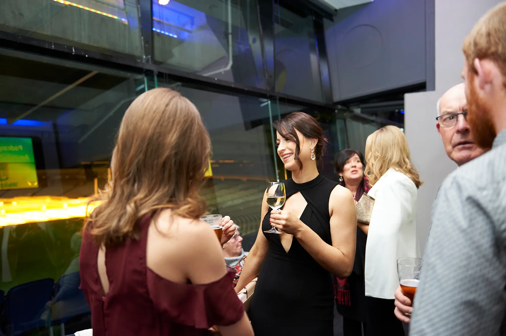 padraig pearses gac awards night mcg guests arrival burgundy black dress Two women engage in conversation during the arrival period of the Padraig Pearses GAC Awards Night & Gala Dinner at the MCG. One wears a burgundy off-shoulder dress holding a beer glass, while the other wears a black keyhole-neck dress with a wine glass, smiling warmly. Guests, including an older man with glasses, mingle in the sleek, modern event space featuring floor-to-ceiling glass windows and blue ambient lighting. The scene captures a sophisticated, welcoming atmosphere as formally dressed attendees socialize, enjoy drinks, and anticipate the evening’s gala festivities.