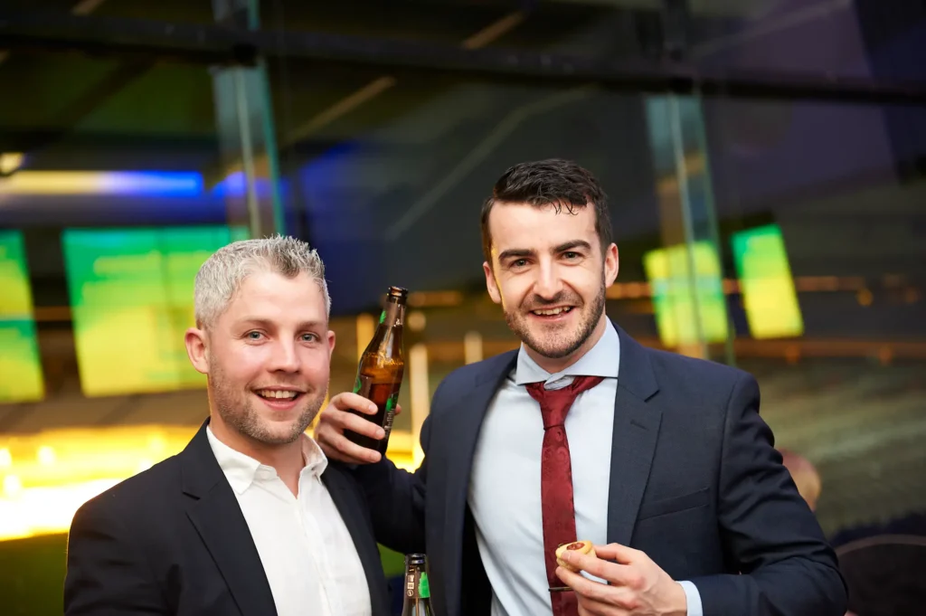 padraig pearses gac awards night mcg guests arrival camaraderie glass wall Two men in dark suits smile warmly at the camera during the arrival period of the Padraig Pearses GAC Awards Night & Gala Dinner at the MCG. One man with grey hair rests his arm on the shoulder of his companion, while the other, wearing a red tie, holds a beer and a small appetizer. Blue and green ambient lighting reflects on glass partitions in the modern event space, and other guests mingle in the blurred background. The scene captures a welcoming, relaxed, and sociable atmosphere as attendees arrive and engage before the main celebrations begin.