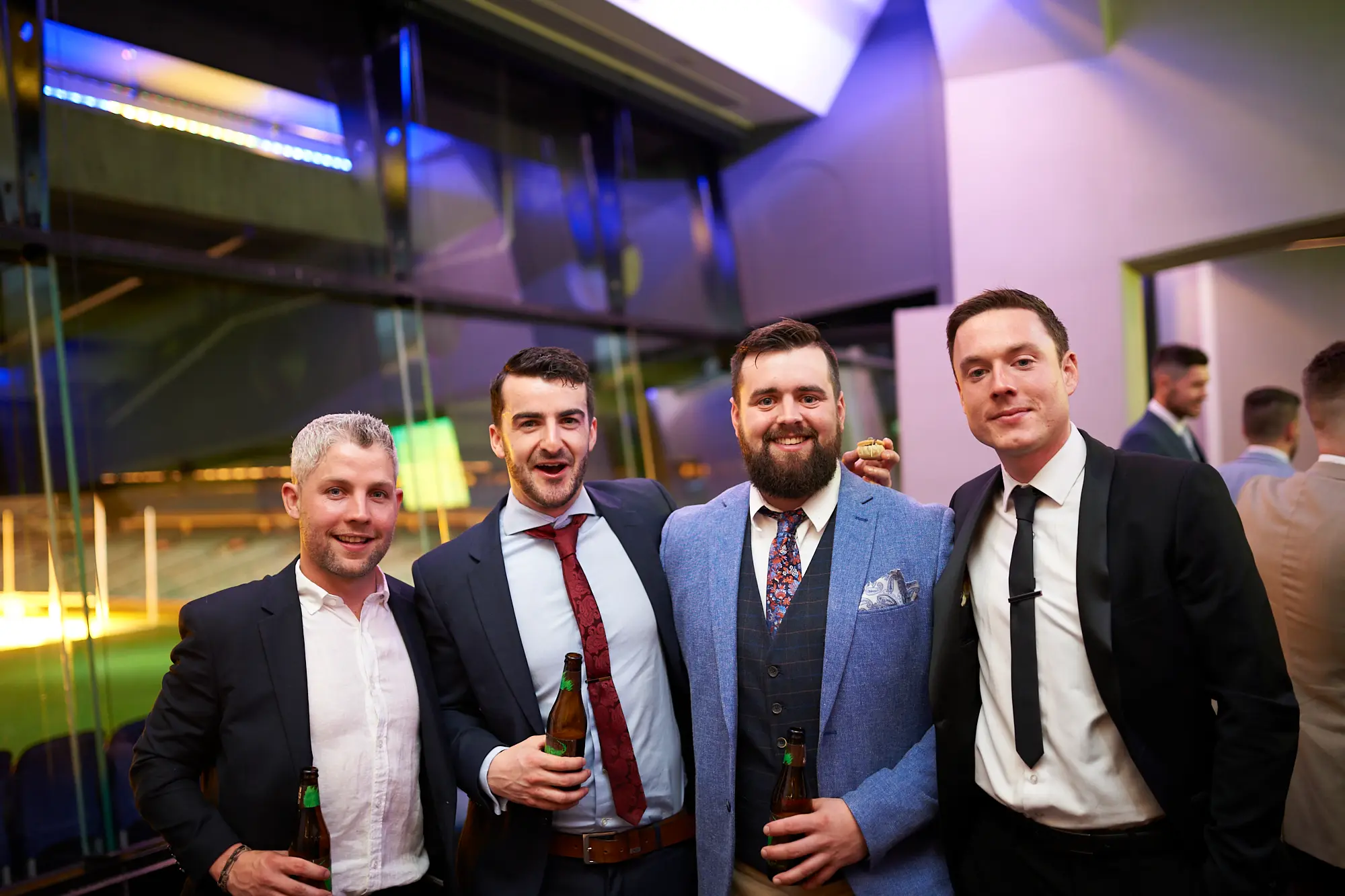 padraig pearses gac awards night mcg guests arrival camaraderie Four men in formal attire share a cheerful moment at the Padraig Pearses GAC Awards Night & Gala Dinner at the MCG, smiling and holding beer bottles as they pose for a photograph. Their close proximity and relaxed expressions convey camaraderie and excitement during the evening’s arrival. Large glass windows reveal blue stadium seating and the dimly lit MCG field beyond, while other guests mingle in the background. The cool, blue-toned lighting highlights the sophisticated yet social atmosphere of the event, capturing the beginning of a memorable and celebratory night.