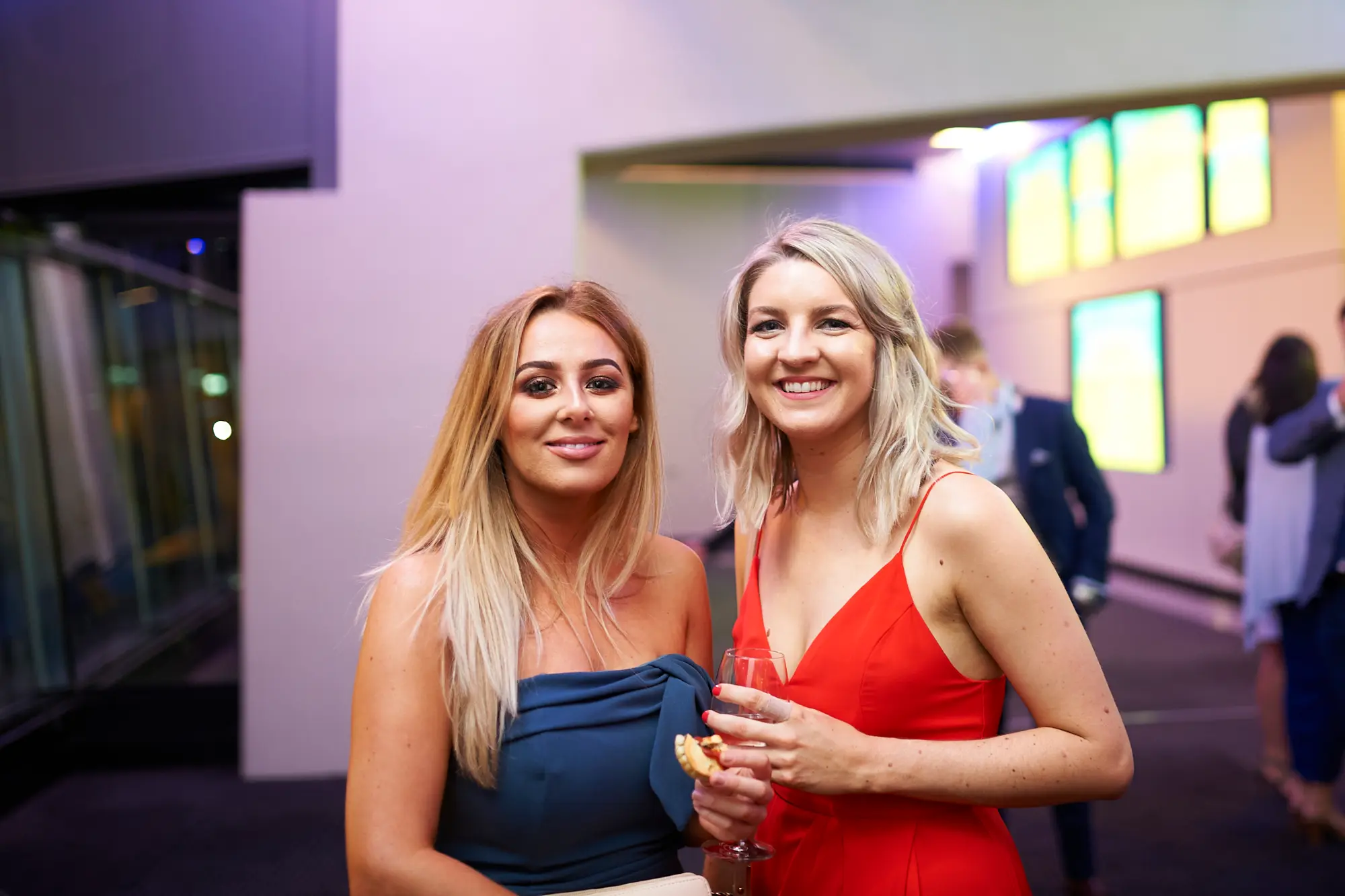 padraig pearses gac awards night mcg guests arrival elegant socializing Two women pose with warm smiles during the arrival period of the Padraig Pearses GAC Awards Night & Gala Dinner at the MCG. The woman in a dark blue strapless dress and the woman in a vibrant red dress holding a wine glass enjoy the social setting. Modern interior design with geometric lines, vertical lit panels, and sleek dark flooring provides a sophisticated backdrop, while other formally dressed guests move and mingle in the background. The scene captures an elegant, welcoming atmosphere as attendees arrive, socialize, and begin the evening’s gala festivities.