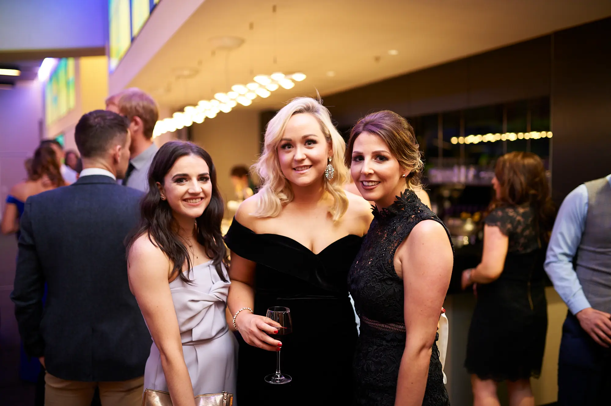 padraig pearses gac awards night mcg guests arrival elegant women black gray lace Three women pose together with warm smiles during the arrival period of the Padraig Pearses GAC Awards Night & Gala Dinner at the MCG. The central woman wears a black velvet off-shoulder dress and holds a wine glass, flanked by women in a light gray twisted bodice dress and a black high-neck lace dress. Their close, friendly poses convey camaraderie and enjoyment. The modern event space features warm spot lighting, vertical panels with blue and green accents, and dark walls and flooring. The scene captures a sophisticated, welcoming atmosphere as guests mingle and prepare for the evening’s gala festivities.