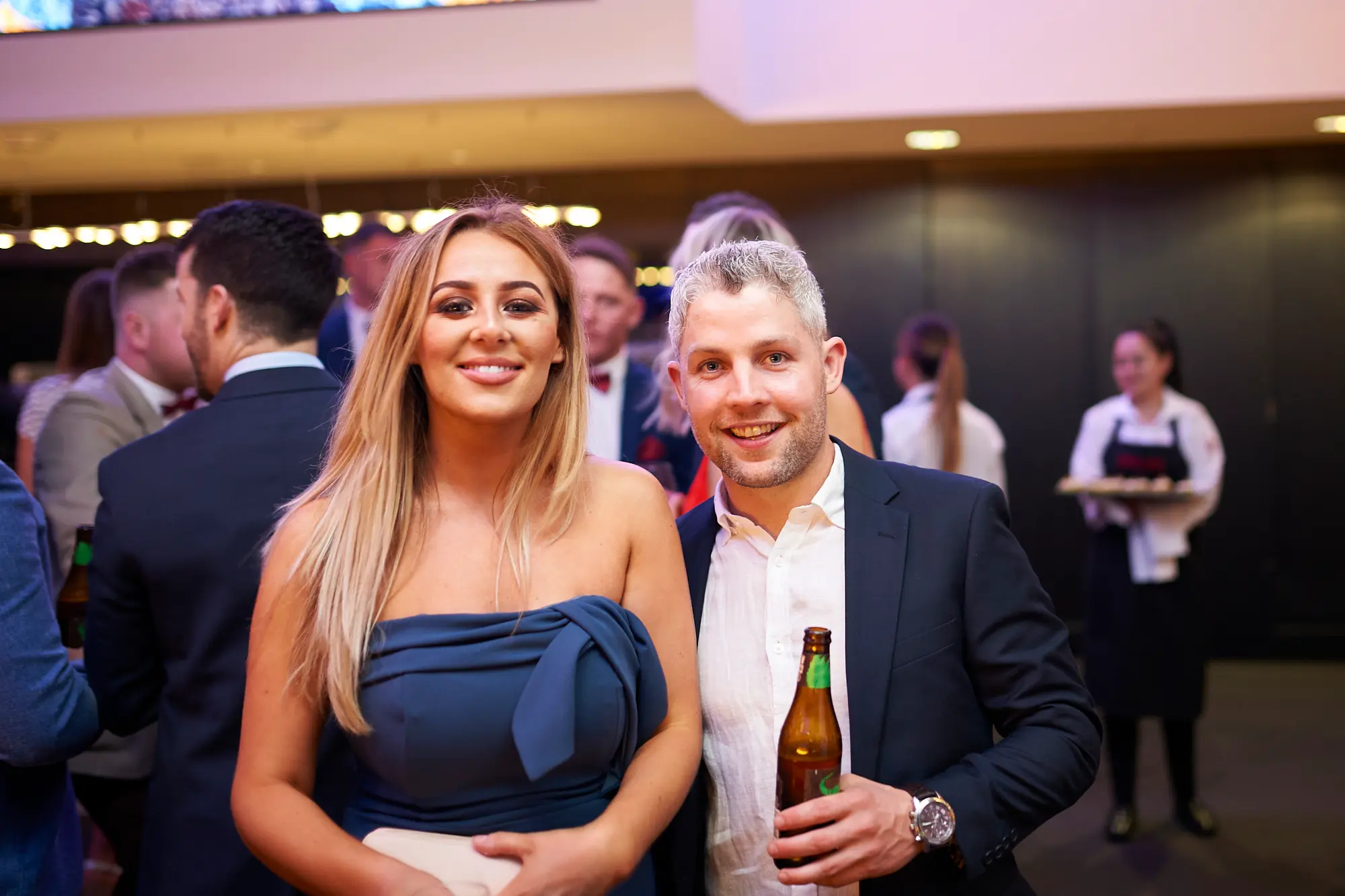 padraig pearses gac awards night mcg guests arrival elegant A woman in a blue strapless dress and a man in a dark suit smile at the camera during the arrival period of the Padraig Pearses GAC Awards Night & Gala Dinner at the MCG. The man holds a beer bottle while the woman carries a light-colored clutch, reflecting a relaxed, social mood. Other formally dressed guests mingle in the dimly lit background, and a staff member with a tray indicates catering service. The scene captures the welcoming, festive, and elegant atmosphere as attendees arrive and engage before the main event begins.