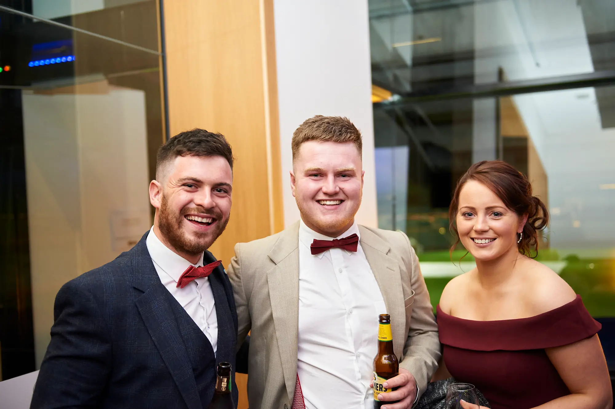 padraig pearses gac awards night mcg guests arrival festive Three guests pose with broad smiles during the arrival of the Padraig Pearses GAC Awards Night & Gala Dinner at the MCG. Two men in suits and red bow ties hold beer bottles, while a woman in a burgundy off-shoulder dress smiles warmly. Their close, relaxed postures reflect friendship and enjoyment. The modern indoor event space features architectural glass panels, sleek walls, and ambient lighting with subtle blue accents. The scene captures a festive, welcoming, and elegant atmosphere as attendees socialize, enjoy drinks, and anticipate the evening’s gala celebrations.