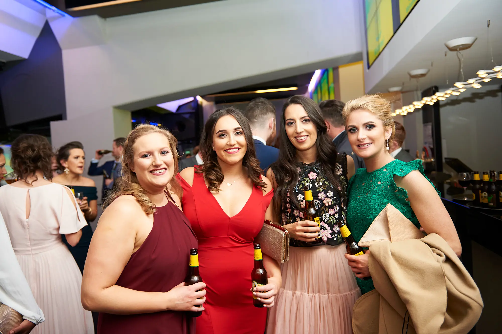padraig pearses gac awards night mcg guests arrival four women beer Four women pose together with warm smiles during the arrival of the Padraig Pearses GAC Awards Night & Gala Dinner at the MCG. Each holds a brown glass beer bottle, enjoying drinks and socializing. One woman carries a beige coat over her arm. The reception area features a long bar stocked with drinks, warm and subtle blue ambient lighting, and sleek modern architecture. Other formally dressed guests mingle in the blurred background. The scene captures a festive, welcoming, and sophisticated atmosphere as attendees arrive, engage socially, and anticipate the evening’s gala celebrations.