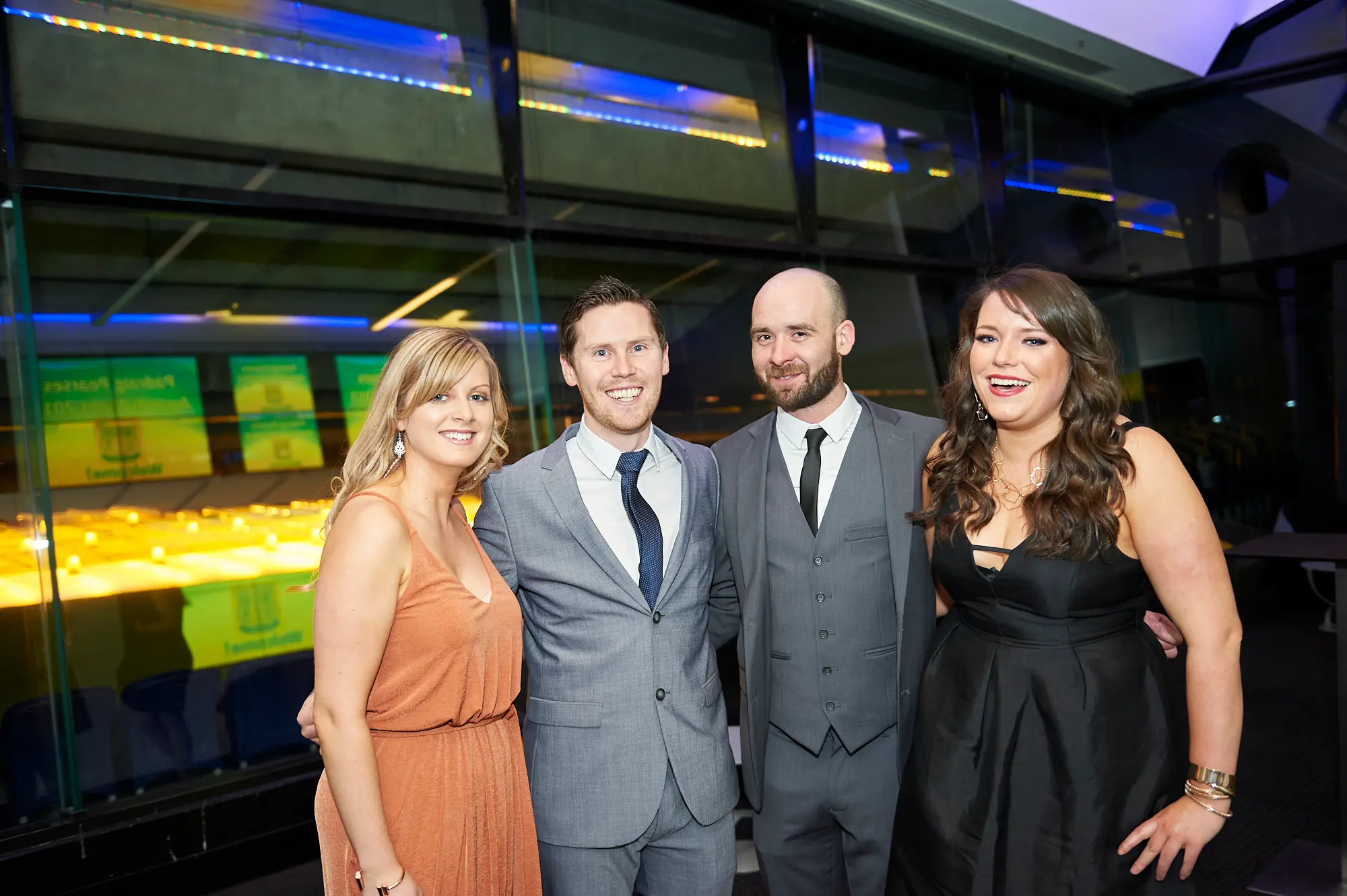 padraig pearses gac awards night mcg guests arrival orange peach black dress Four guests pose warmly during the arrival period of the Padraig Pearses GAC Awards Night & Gala Dinner at the MCG. Two men in grey suits and ties smile broadly, while a woman in an orange-peach dress and another in a black evening gown engage enthusiastically with the camera. Floor-to-ceiling glass windows reveal dark stadium seating, and vibrant blue and yellow-green lighting accents add a modern, dynamic feel. The scene captures a sophisticated, welcoming atmosphere as formally dressed attendees arrive, socialize, and anticipate the evening’s gala celebrations.
