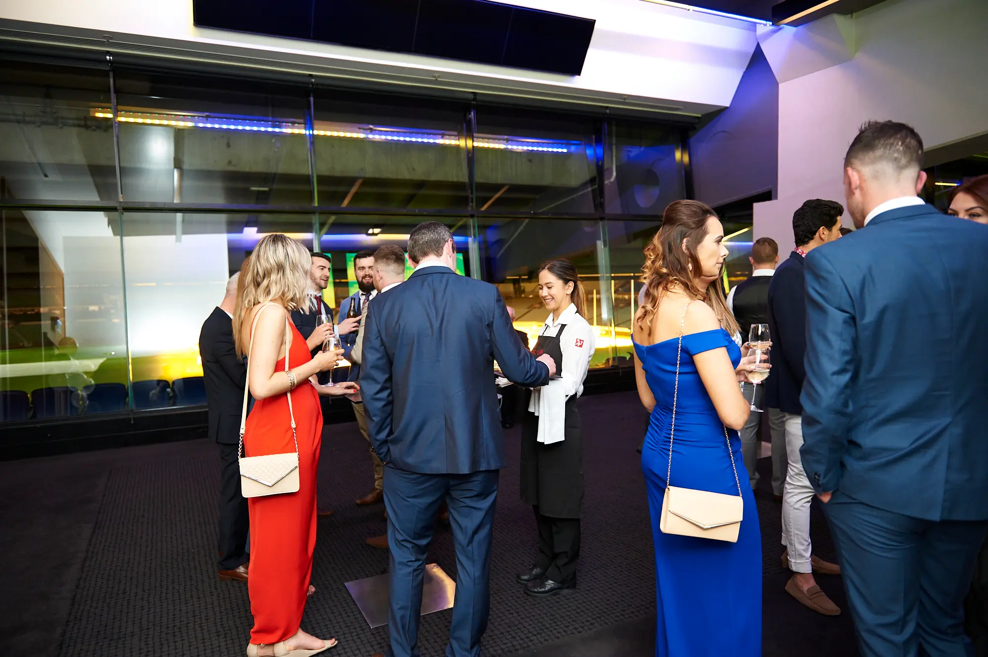 padraig pearses gac awards night mcg guests arrival socializing Guests mingle during the arrival period of the Padraig Pearses GAC Awards Night & Gala Dinner at the MCG. A female staff member in a white shirt and black apron shakes hands with a male guest in a blue suit, while women in a blue off-shoulder gown and red dress hold drinks and socialize. Large glass windows reveal dimly lit stadium seating and the MCG field, with blue neon and warm interior lighting enhancing the sophisticated atmosphere. The scene captures elegant social interactions and anticipation as attendees enjoy drinks and conversation before the main event.