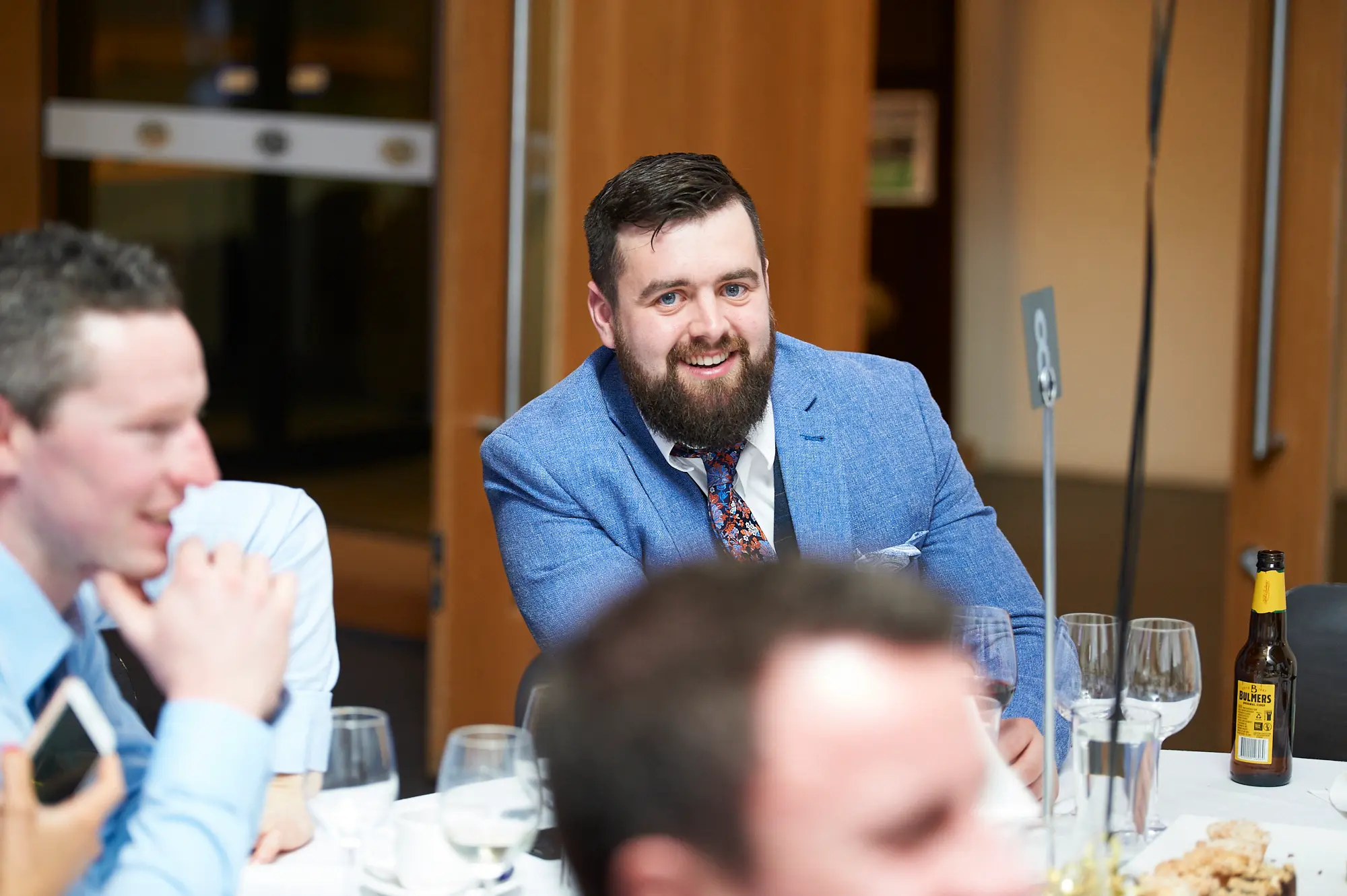 padraig pearses gac awards night mcg guests dinner table blue blazer beard Guests share a relaxed moment of socializing at a dinner table during the Padraig Pearses GAC Awards Night & Gala Dinner at the MCG. A man with a dark beard in a light blue blazer smiles warmly at the camera, while another man in a light blue shirt engages in conversation nearby. The white-clothed table features wine glasses, plates, and a brown glass beer bottle. Modern wooden paneling, a glass partition, and warm ambient lighting create a contemporary, formal yet relaxed atmosphere, capturing camaraderie, enjoyment, and friendly interactions among attendees during the gala dinner.