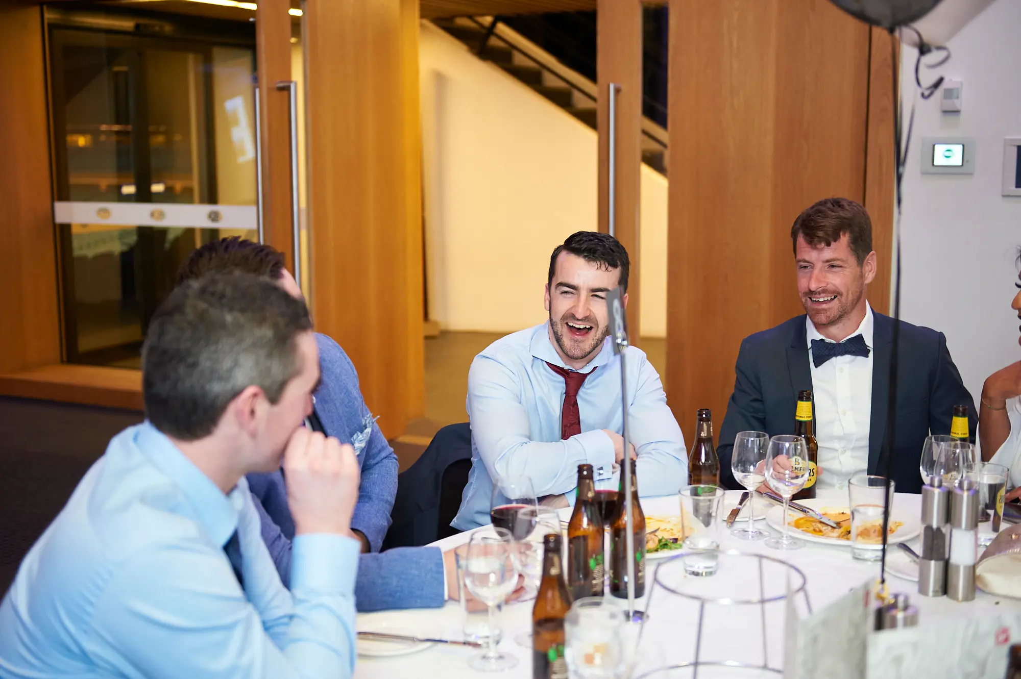 padraig pearses gac awards night mcg guests dinner table blue shirt red tie Guests share a lively moment of conversation and camaraderie at a dinner table during the Padraig Pearses GAC Awards Night & Gala Dinner at the MCG. A man in a light blue shirt with a red tie laughs heartily, leaning forward, while a man in a navy suit with a patterned bow tie smiles broadly holding a beer bottle. Another man in the foreground looks engaged, reacting to the conversation. The white-clothed table features wine glasses, cutlery, plates, and beer bottles. The scene conveys a festive, social, and relaxed atmosphere as attendees enjoy dinner, drinks, and lively interaction during the gala event.
