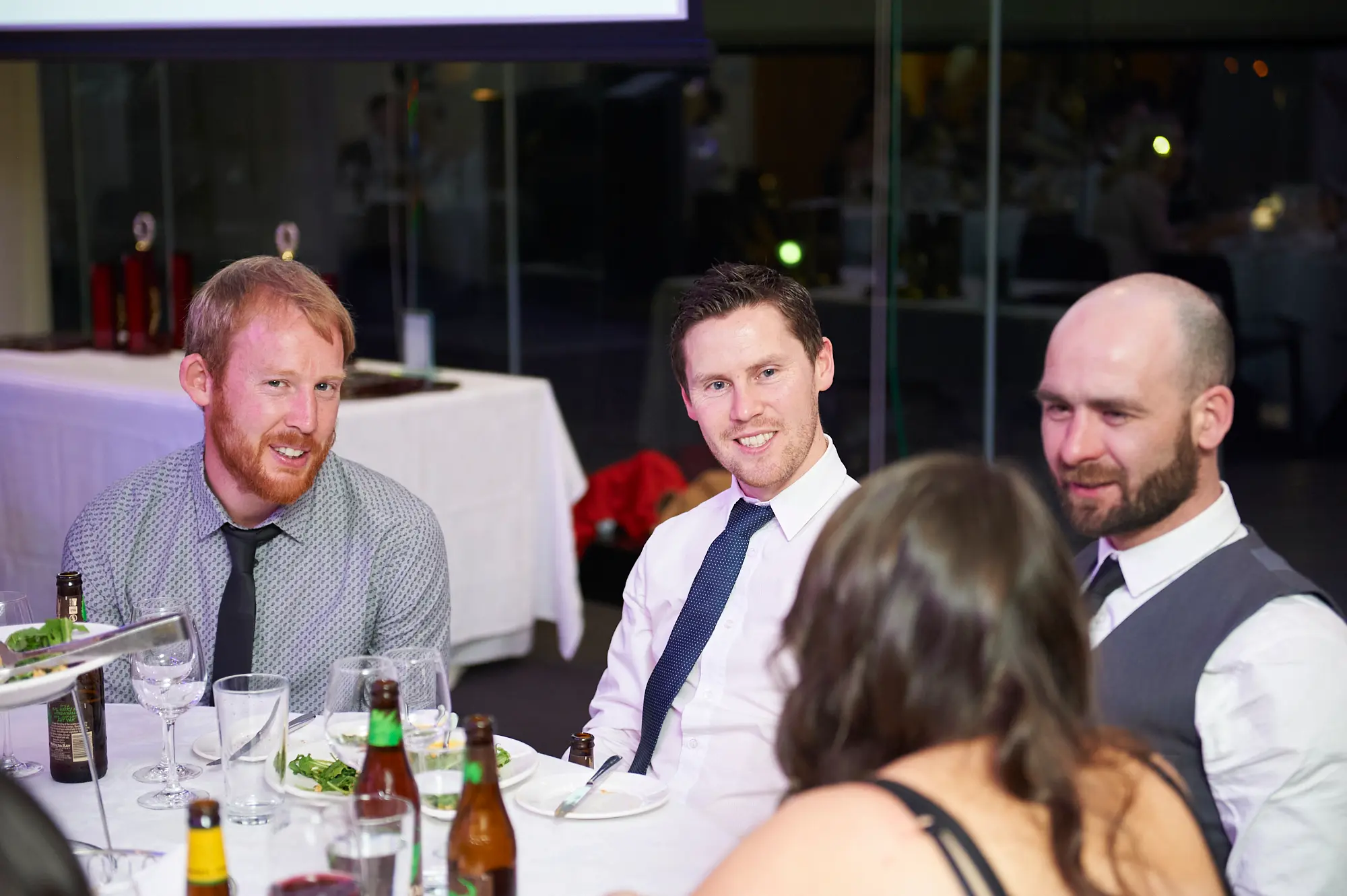 padraig pearses gac awards night mcg guests dinner table camaraderie Three men and one woman share a relaxed moment at a dinner table during the Padraig Pearses GAC Awards Night & Gala Dinner at the MCG. Two men smile warmly at the camera, while a bald man with a dark beard looks down, and the woman with her back to the camera engages in conversation. The table is set with wine glasses, plates, cutlery, and beer bottles. Dim lighting, glass partitions, and warm ambient lights in the background create a formal yet intimate and social atmosphere, capturing camaraderie, conversation, and enjoyment during the gala dinner.