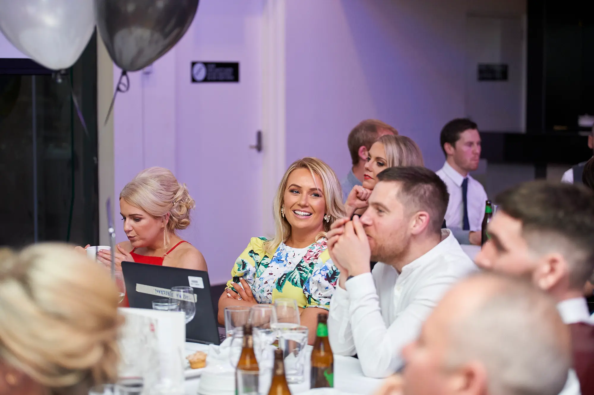 padraig pearses gac awards night mcg guests dinner table floral red laptop Guests enjoy a lively dining moment during the Padraig Pearses GAC Awards Night & Gala Dinner at the MCG. A woman in a floral dress smiles warmly at the camera, while a man next to her leans forward on the table engaged in conversation, and a woman in a red dress focuses on a laptop in front of her. The white-clothed table features wine glasses, cutlery, and beer bottles. White and gray/black balloons and a festive indoor hall set a celebratory, social, and relaxed atmosphere as attendees enjoy dinner, drinks, and engaging interaction during the gala event.
