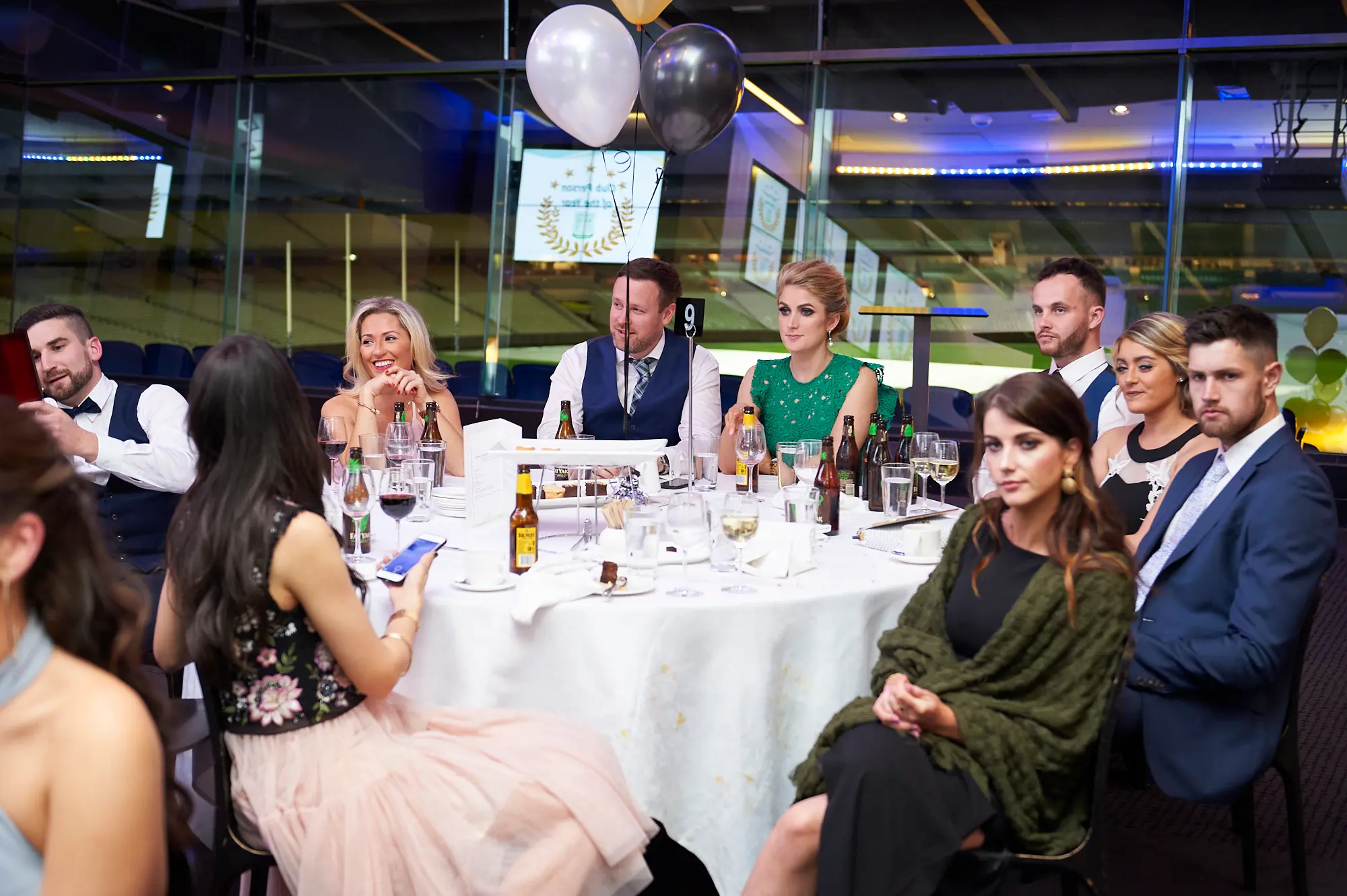 padraig pearses gac awards night mcg guests dinner table green dress Guests enjoy a lively dining moment at a white-clothed round table during the Padraig Pearses GAC Awards Night & Gala Dinner at the MCG. A woman in a green dress smiles broadly, a man in a navy waistcoat holds a beer bottle, and a woman in a pink skirt and black top takes a photo. Another woman in a black dress with an olive-green shawl looks poised at the camera. The table features wine glasses, plates, cutlery, and beer bottles, while balloons, large glass windows, and blue ambient lighting create a festive, social, and celebratory atmosphere during the gala event.