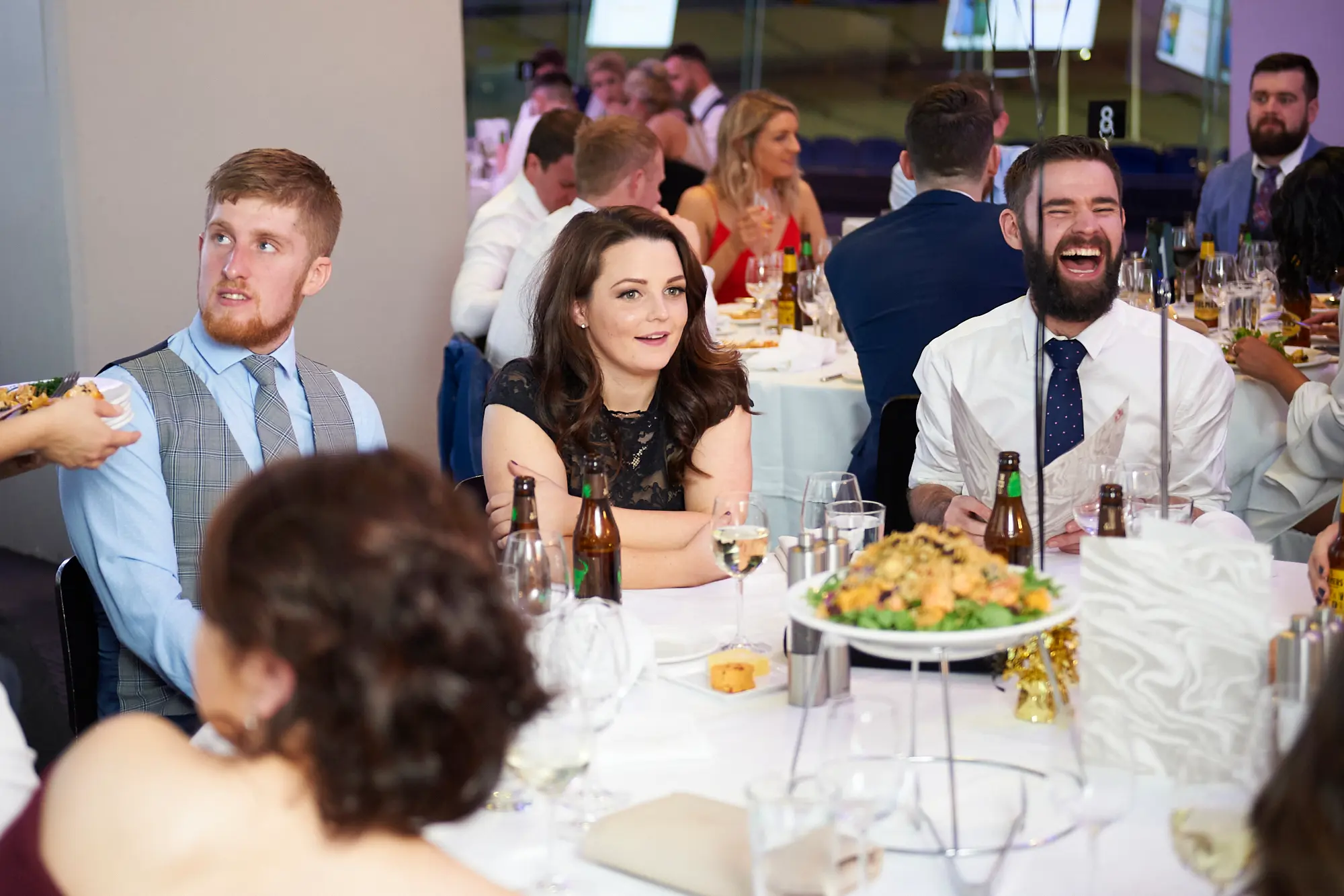 padraig pearses gac awards night mcg guests dinner table laughter Guests share a lively dining moment during the Padraig Pearses GAC Awards Night & Gala Dinner at the MCG. A man in a white shirt and dark blue tie laughs heartily holding a beer bottle, a woman in a black lace dress smiles warmly, and a man in a light blue shirt with grey checkered waistcoat looks on with interest. The table features wine glasses, cutlery, and beer bottles. White-clothed tables, elegantly dressed guests, and warm ambient lighting create a festive, social, and celebratory atmosphere as attendees enjoy dinner, drinks, and camaraderie in the large gala venue.