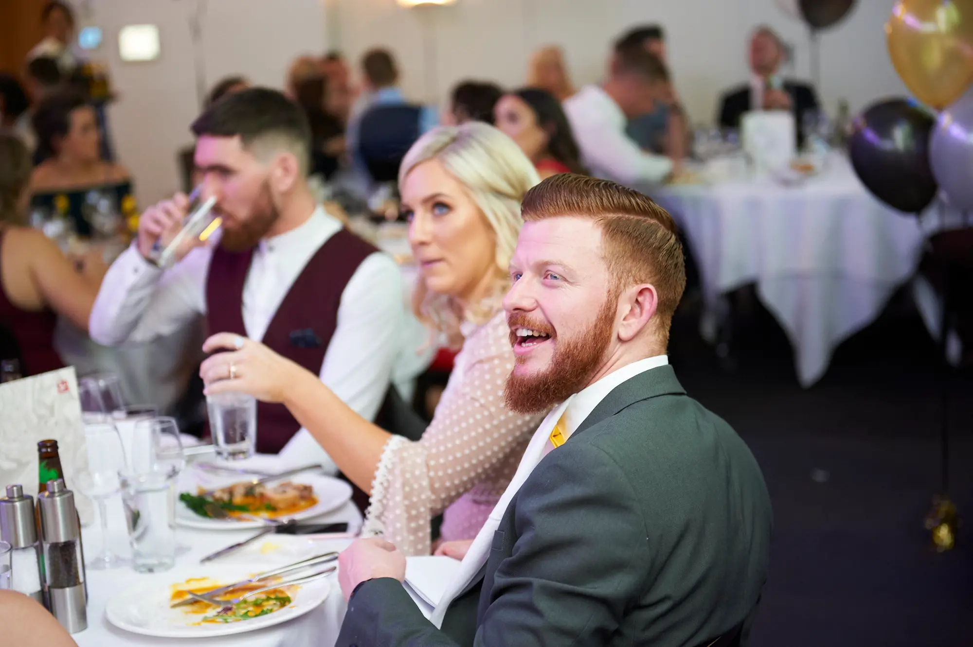padraig pearses gac awards night mcg guests dinner table polka dot maroon Three guests enjoy a lively dining moment during the Padraig Pearses GAC Awards Night & Gala Dinner at the MCG. A man in a maroon waistcoat drinks from a glass, a woman in a white polka-dot top looks engaged holding water, and a man in a dark grey suit with a yellow tie smiles broadly. White-clothed round tables, black and gold balloons, and warmly lit surroundings set a festive and social atmosphere. The scene captures formal yet lively engagement as guests enjoy dinner, drinks, and friendly conversation in a large, elegant function room.