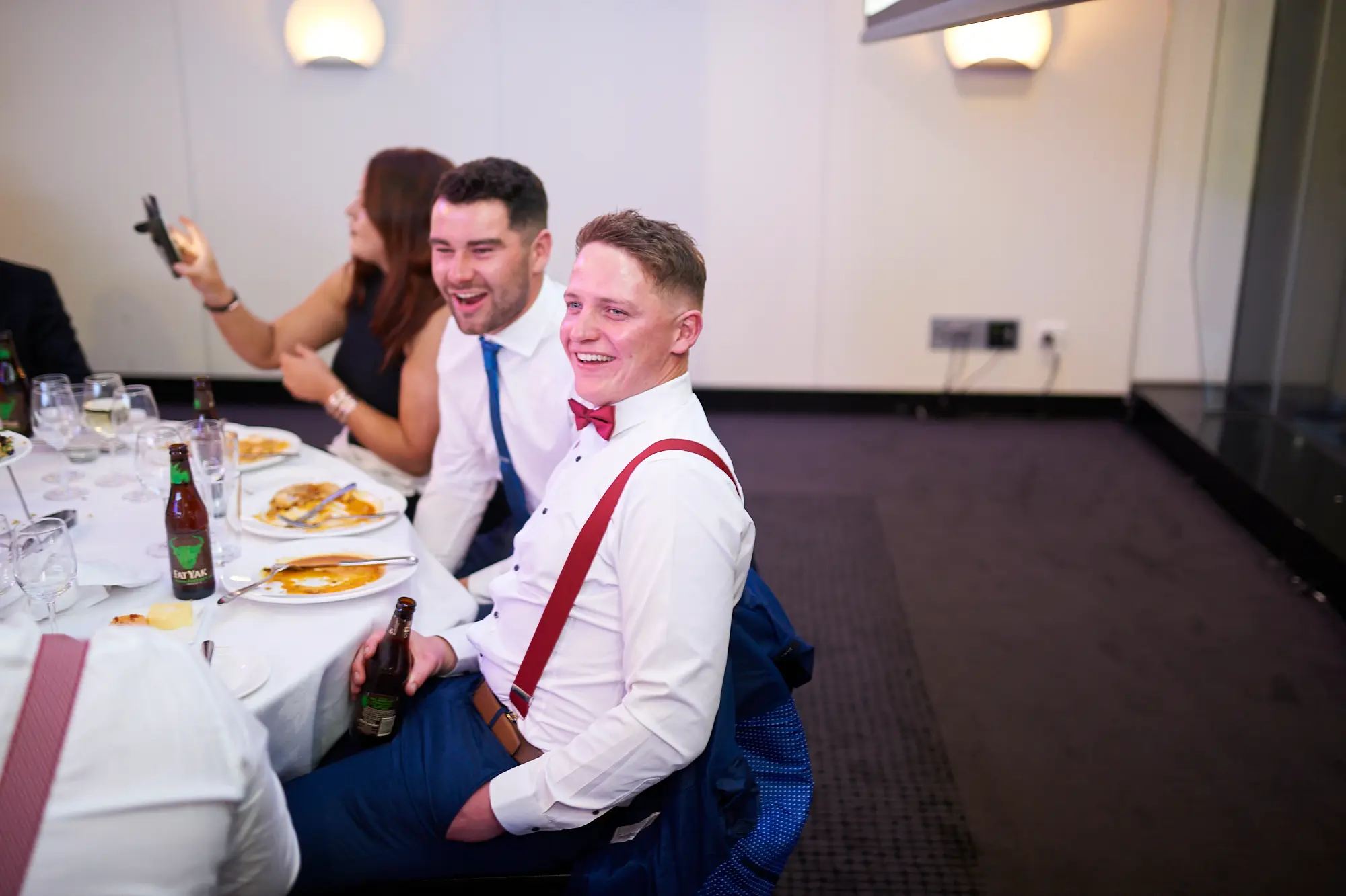 padraig pearses gac awards night mcg guests dinner table red bow tie Guests share a lively and joyful moment at a dinner table during the Padraig Pearses GAC Awards Night & Gala Dinner at the MCG. A man in a white shirt, red bow tie, and red suspenders laughs heartily while holding a beer bottle, seated next to a man in a blue tie who is also laughing. A woman in the background holds up her phone, capturing the moment. The table features white tablecloths, plates, glasses, and bottles. The scene conveys a festive, social, and relaxed atmosphere as attendees enjoy dinner, drinks, and camaraderie during the gala event.