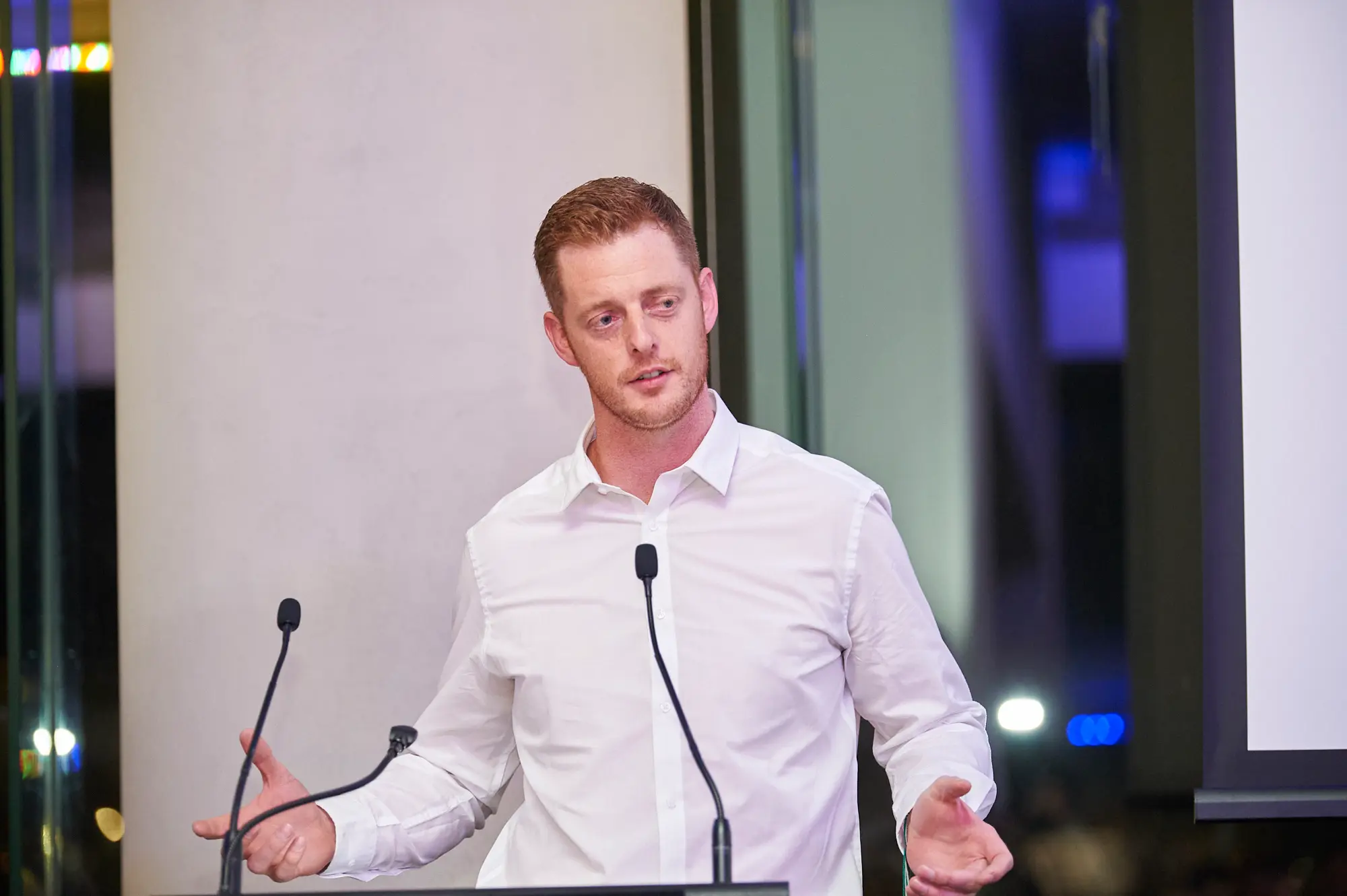 padraig pearses gac awards night mcg host podium speaking A reddish-blonde man in a white collared shirt stands behind a black lectern, speaking animatedly into two microphones at the Padraig Pearses GAC Awards Night at the MCG. His hands gesture outward as he engages the audience. A large white screen and modern glass partitions with blue and purple ambient lighting create a sophisticated backdrop, highlighting the professional and lively atmosphere of the gala dinner presentation.