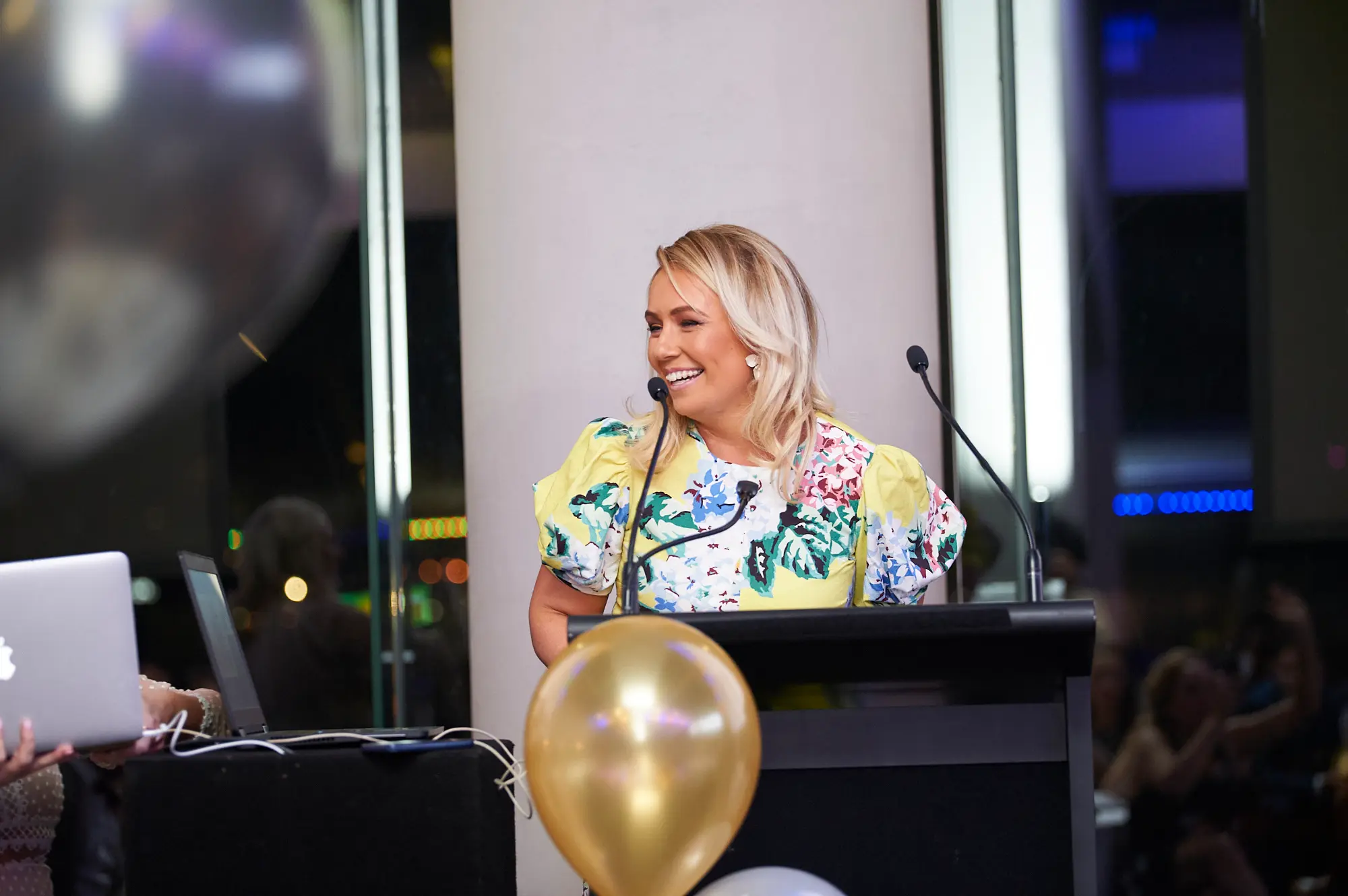 padraig pearses gac awards night mcg hosting podium yellow dress A blonde woman in a bright yellow floral dress stands behind a black lectern during the Pearses Awards Night at the MCG, smiling broadly and engaging with the audience. A gold balloon is in the foreground, and a dark silver balloon is visible on the left. An Apple laptop sits nearby. Glass partitions reveal seated guests in the dimly lit background with blue accent lighting. Her confident posture and joyful expression convey a lively, professional, and celebratory atmosphere, highlighting an engaging moment of hosting and presentation at the gala dinner.