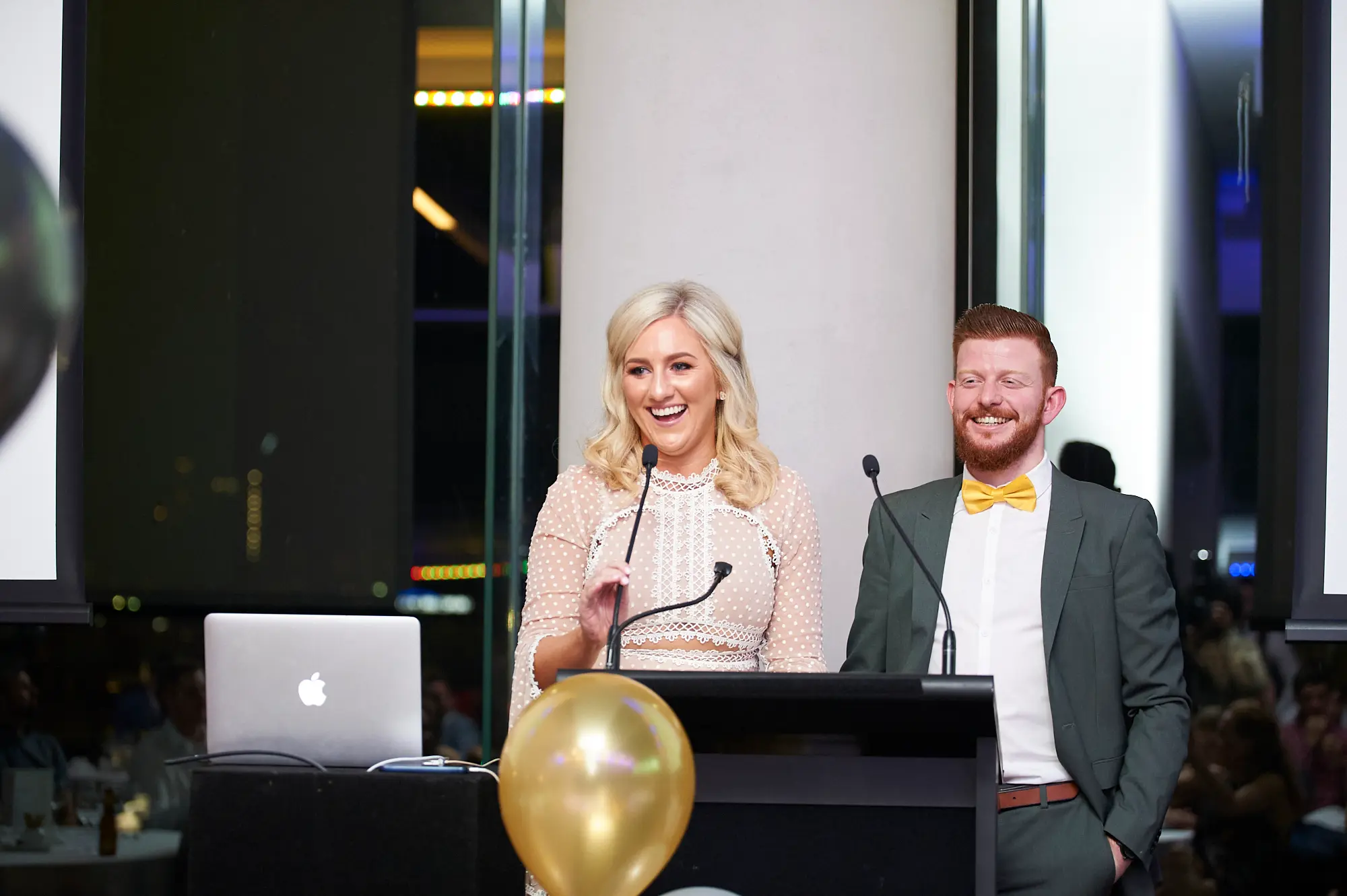 padraig pearses gac awards night mcg hosts podium presentation Two hosts stand behind a black lectern during the Padraig Pearses GAC Awards Night at the MCG. The woman holds a microphone, smiling and engaging the audience, while the man stands relaxed with his hands in his pockets. A gold balloon and an Apple laptop sit on the lectern. Blue and warm yellow accent lighting reflects off sleek columns and glass partitions, while formally dressed guests are visible in the blurred background, creating an elegant, professional, and celebratory atmosphere.