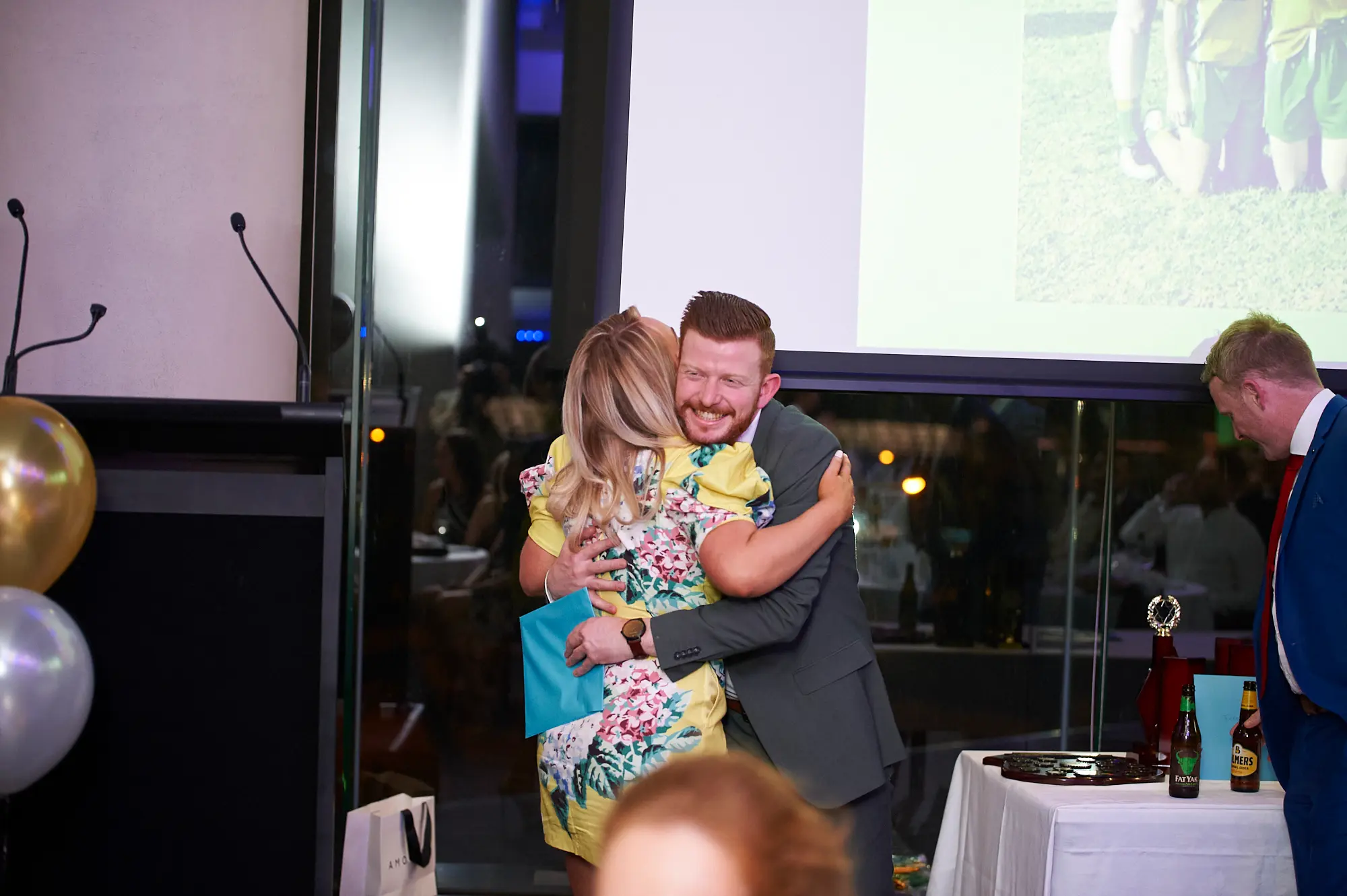padraig pearses gac awards night mcg joyful embrace recognition A man with a red beard in a grey suit warmly embraces a woman in a yellow floral dress holding a blue award at the Pearses Awards Night at the MCG. He smiles broadly at the camera, radiating pride and happiness. Gold and silver balloons, a small trophy, and a large projection screen showing a blurred sports image frame the celebratory moment, while other guests sit at tables in the background, highlighting the festive, personal, and engaging atmosphere of this awards presentation.