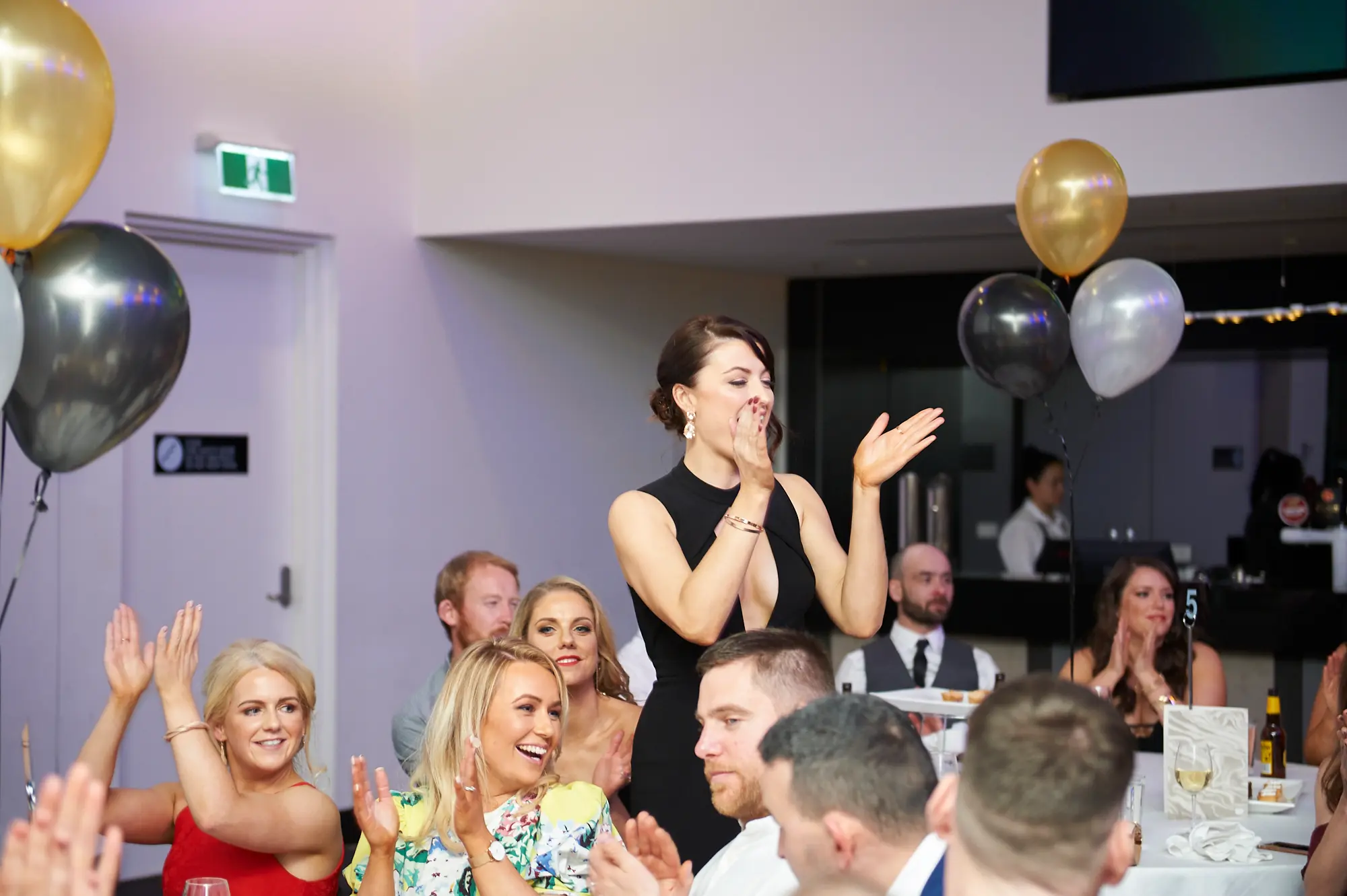 padraig pearses gac awards night mcg lively applause celebration Guests at the Pearses Awards Night at the MCG are clapping and celebrating while seated at round tables. A woman in a black dress smiles broadly with hands raised, while a woman in red and another in floral attire clap and laugh enthusiastically. Gold, silver, and black balloons float around the room, and formal table settings with white tablecloths and wine glasses complete the scene. Other guests in elegant attire are engaged in the celebration, looking toward a stage or presentation, capturing a lively, festive, and joyous moment of recognition and shared excitement during the gala dinner.