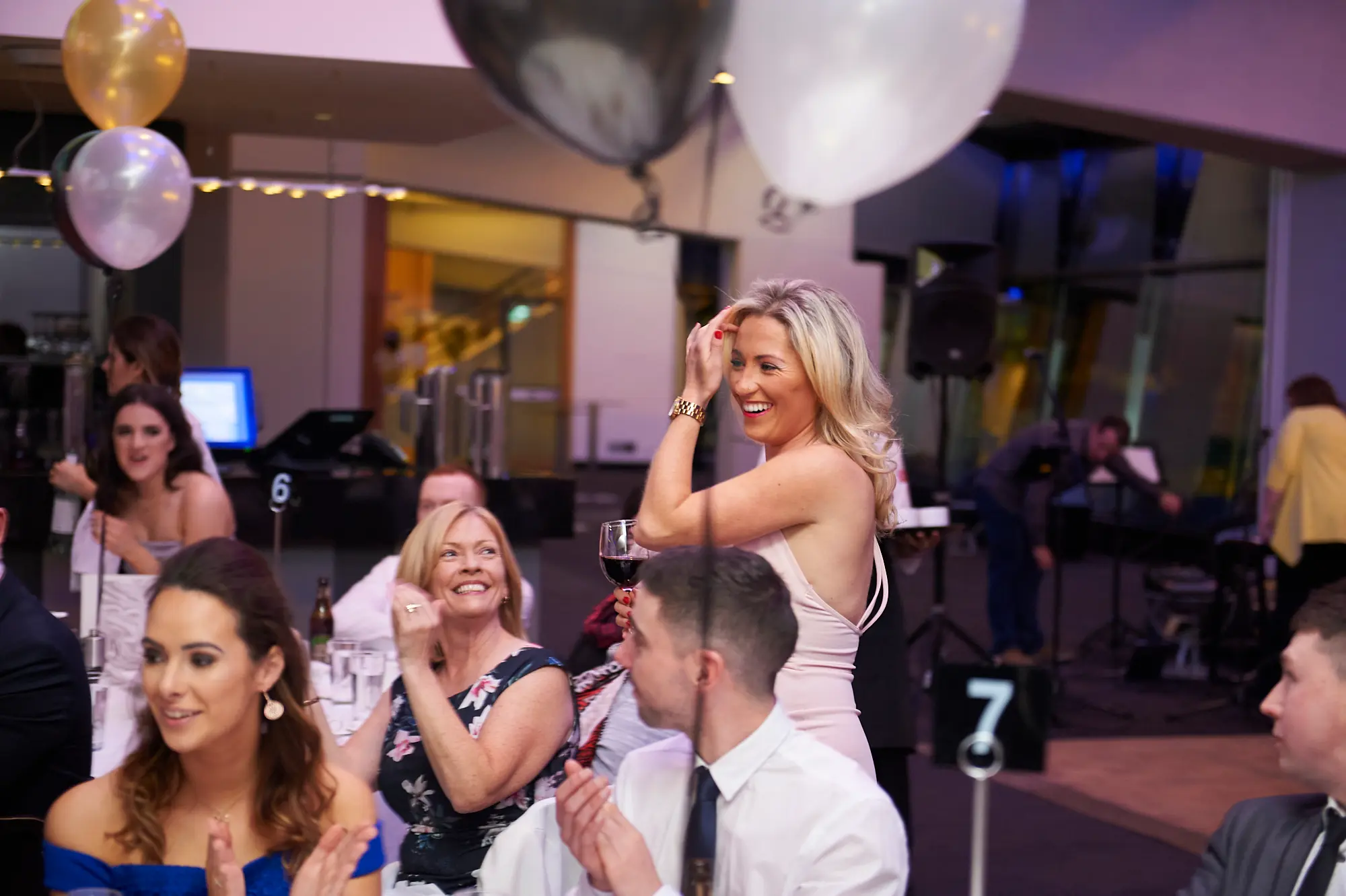 padraig pearses gac awards night mcg lively applause entertainment Guests at the Pearses Awards Night at the MCG are celebrating and applauding while seated at round tables. A woman in a light pink dress raises her hand to her hair and holds a wine glass, smiling broadly, while a woman in floral attire laughs heartily. A man in a white shirt claps, and another woman in blue joins in. White tablecloths, wine glasses, and gold, silver, and black balloons surround the tables. In the background, a stage with musical equipment and other formally dressed guests highlights the festive, social, and engaging atmosphere of this lively gala moment.