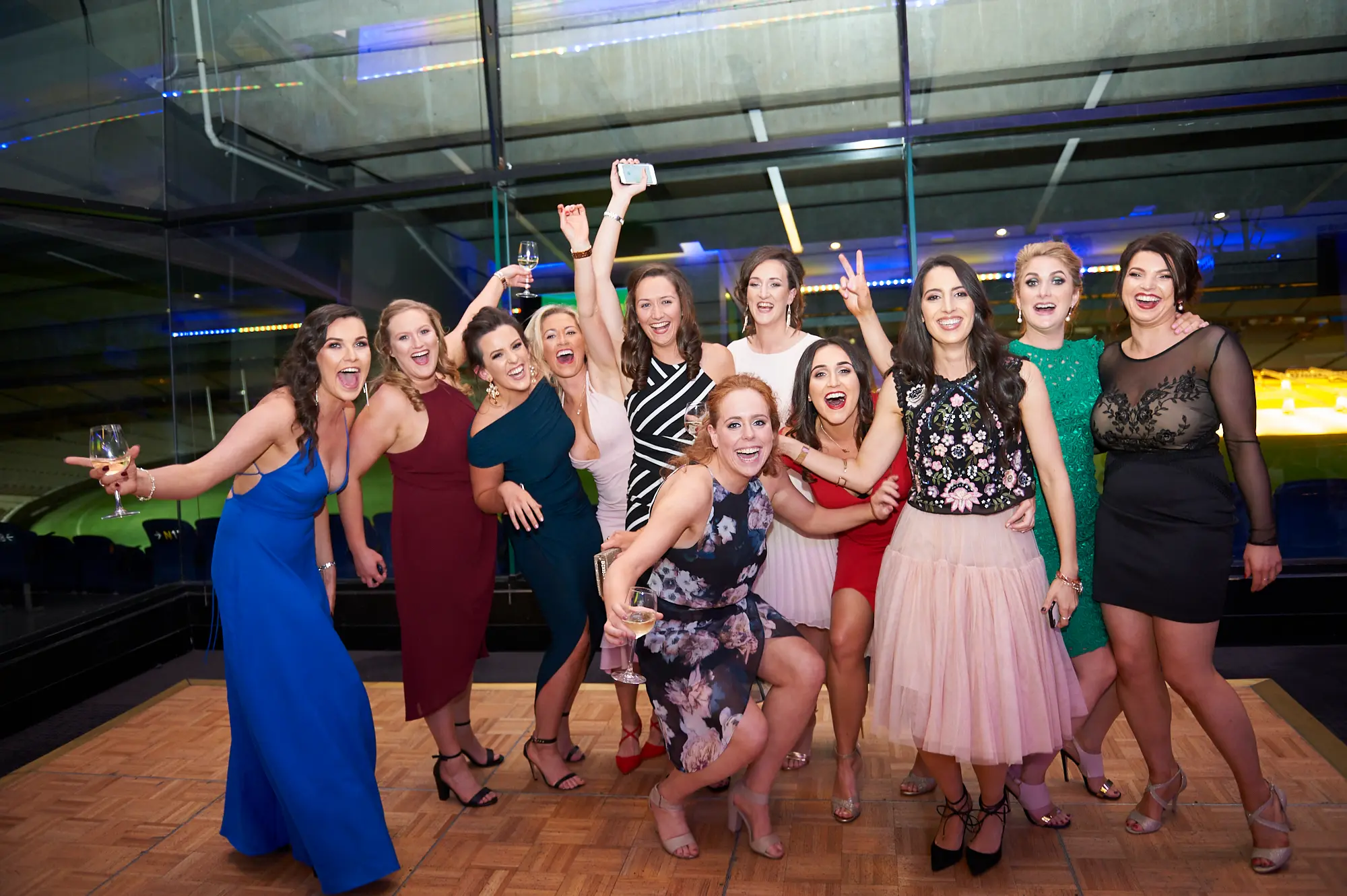 padraig pearses gac awards night mcg lively women celebration dancefloor group A lively group of ten women gather closely on the wooden dance floor during the Pearses Awards Night & Gala Dinner at the MCG, laughing, cheering, and posing with joyful energy. Some raise their arms, others hold wine glasses, crouch playfully, or make peace signs. Behind them, floor-to-ceiling glass panels reveal blue stadium seating and the softly illuminated MCG field, enhanced by vibrant blue accent lighting. The moment captures pure camaraderie, excitement, and the spirited party atmosphere of the evening.