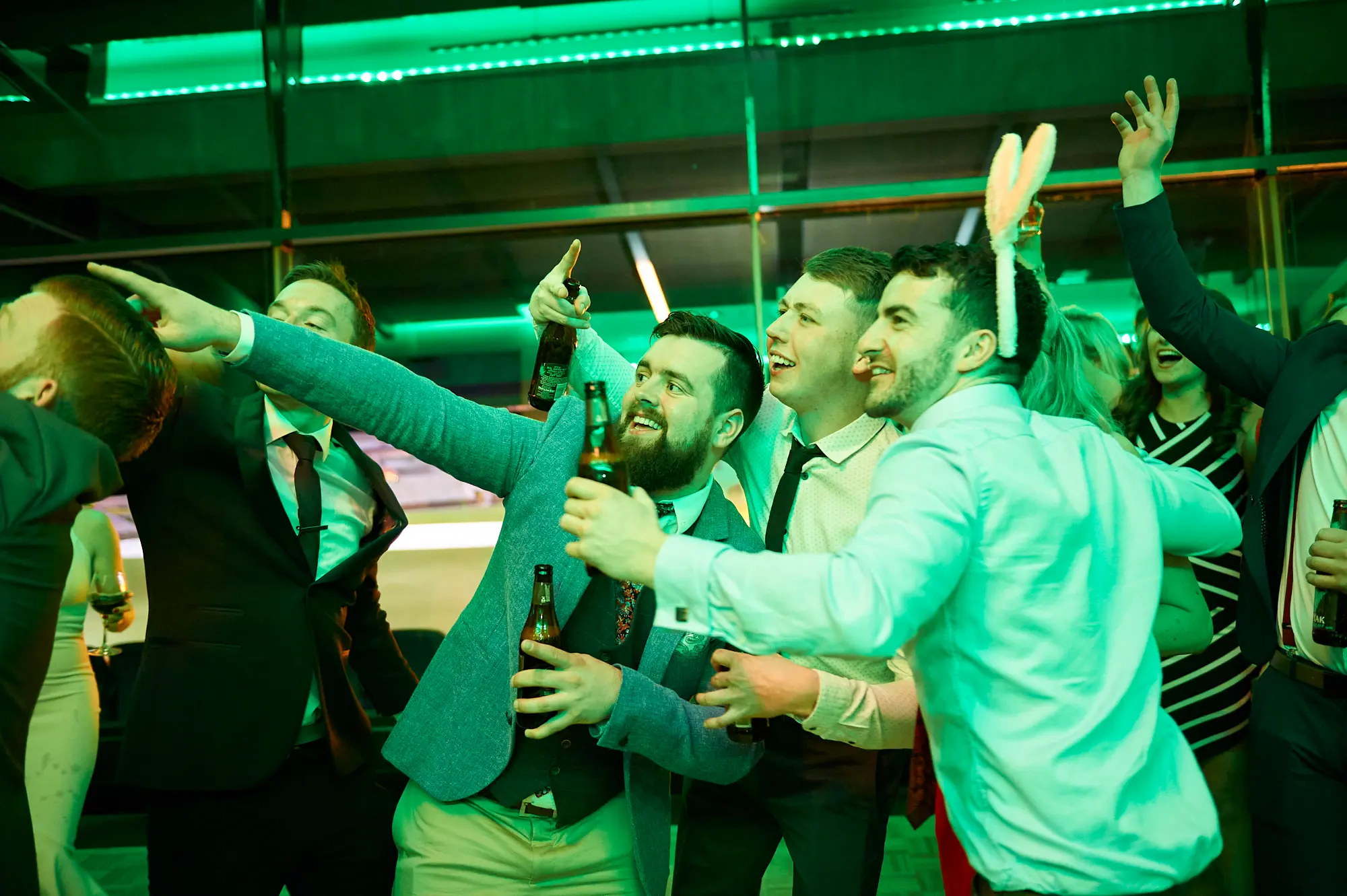 padraig pearses gac awards night mcg party celebration A bearded man in a white shirt and tie celebrates energetically at the Pearses Awards Night at the MCG, holding a beer bottle with a wide-eyed, ecstatic expression. A woman in a black dress cheers beside him, phone raised to capture the moment. Other formally dressed guests join the lively celebration, surrounded by tiered stadium seating and vibrant green ambient lighting. Reflections from glass partitions add depth, highlighting the transition from formal dinner to a spirited party. The image captures a dynamic, joyful, and social atmosphere filled with laughter, excitement, and shared revelry.
