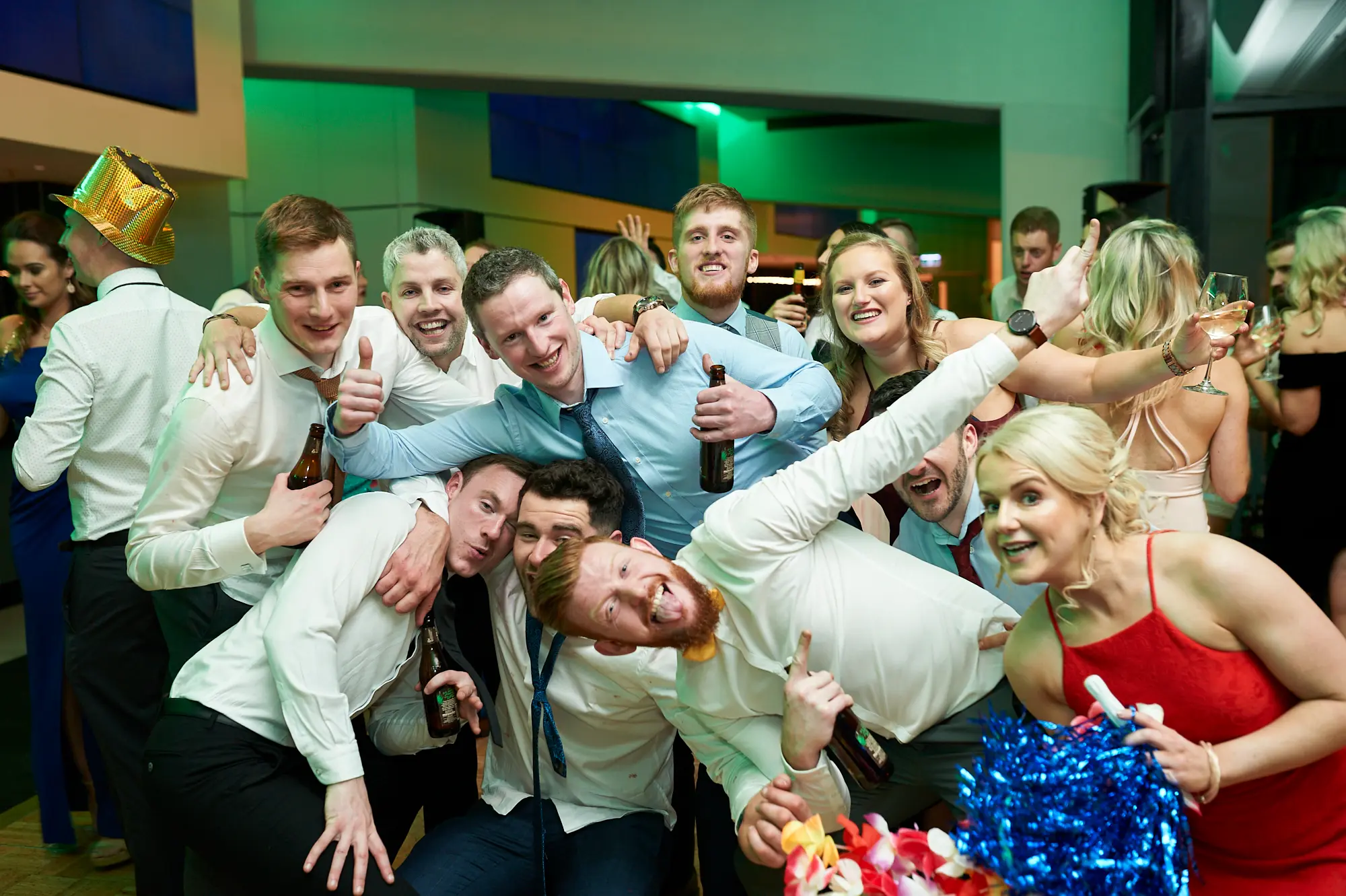 padraig pearses gac awards night mcg party energy Guests celebrate energetically at the Pearses Awards Night at the MCG, with a bearded man in a white shirt and tie gesturing excitedly while holding a beer. A woman in a black dress cheers with her phone raised, capturing the moment. Nearby men in formal wear join the festive mood. The tiered stadium seating and mirrored glass partitions reflect vibrant green ambient lighting, enhancing the dynamic atmosphere. The scene conveys a lively, joyful, and spirited after-party following the formal gala, with guests fully engaged in shared excitement and celebration.