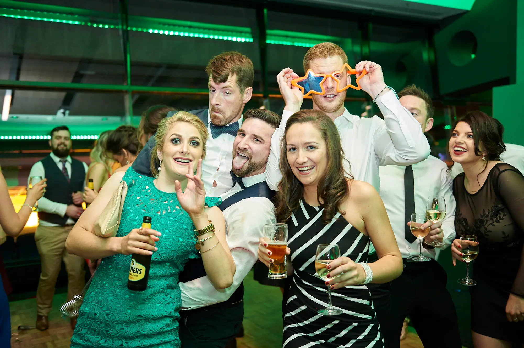padraig pearses gac awards night mcg party fun A lively group of guests enjoys the Pearses Awards Night at the MCG, dressed in formal attire. In the foreground, a woman in a green lace dress waves and holds a beer, while a man next to her in a white shirt and waistcoat playfully sticks out his tongue. Another man wears star-shaped novelty glasses, and others hold wine glasses, laughing and smiling. The modern venue features large glass partitions revealing dark stadium seating, with vibrant green ambient lighting reflecting off the glass. The scene conveys a festive, energetic, and highly social party atmosphere.
