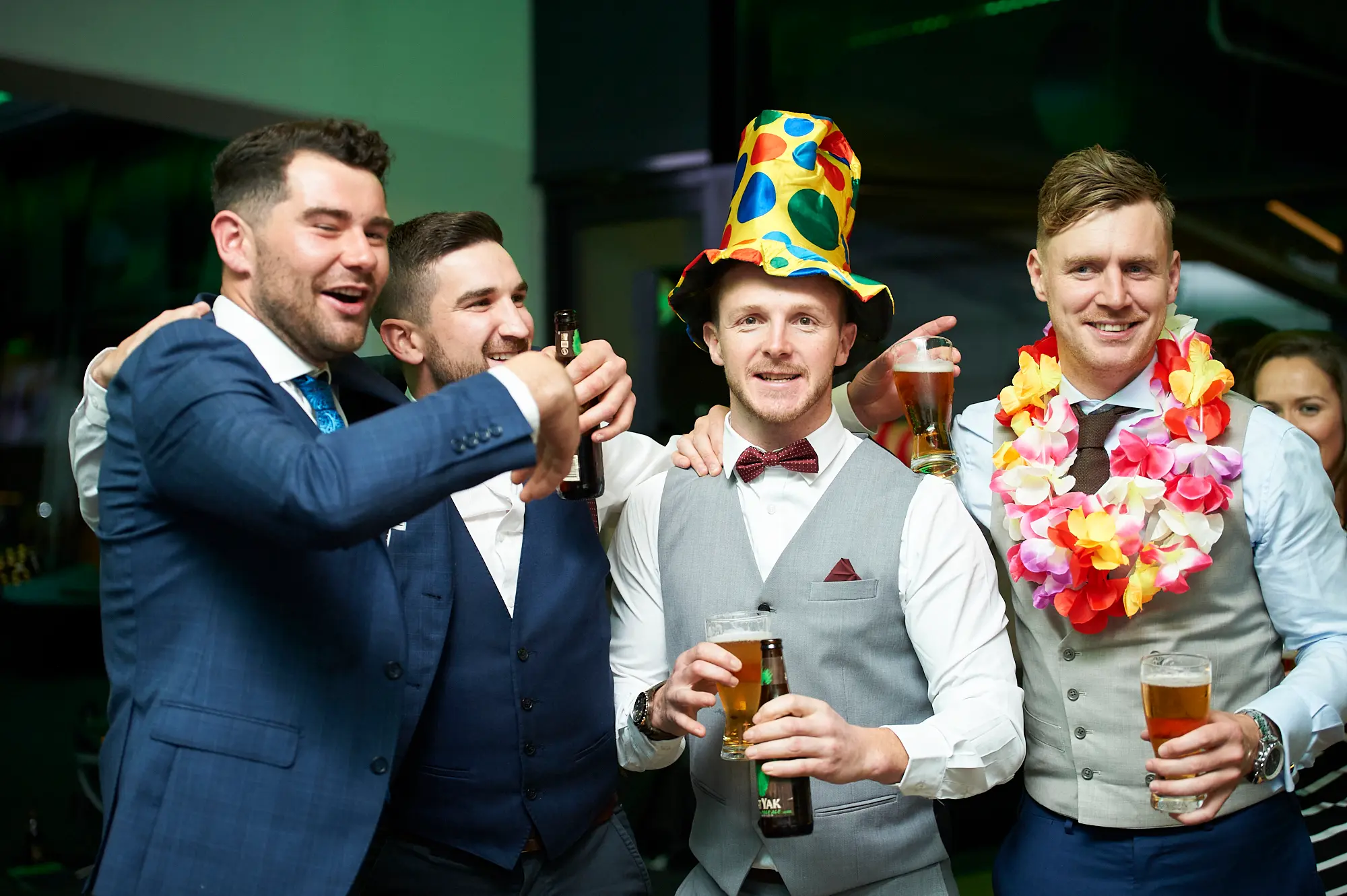 padraig pearses gac awards night mcg party hat toast A lively candid of four men in formal attire celebrating at the Pearses Awards Night & Gala Dinner at the MCG. Two men are clinking beer bottles in a toast, one wears a colorful polka-dot party hat, and another has a bright floral lei. Their broad smiles and relaxed poses convey camaraderie and festive energy, set in a modern indoor event space with green ambient lighting and other guests faintly visible, capturing the fun and social atmosphere of the gala event.