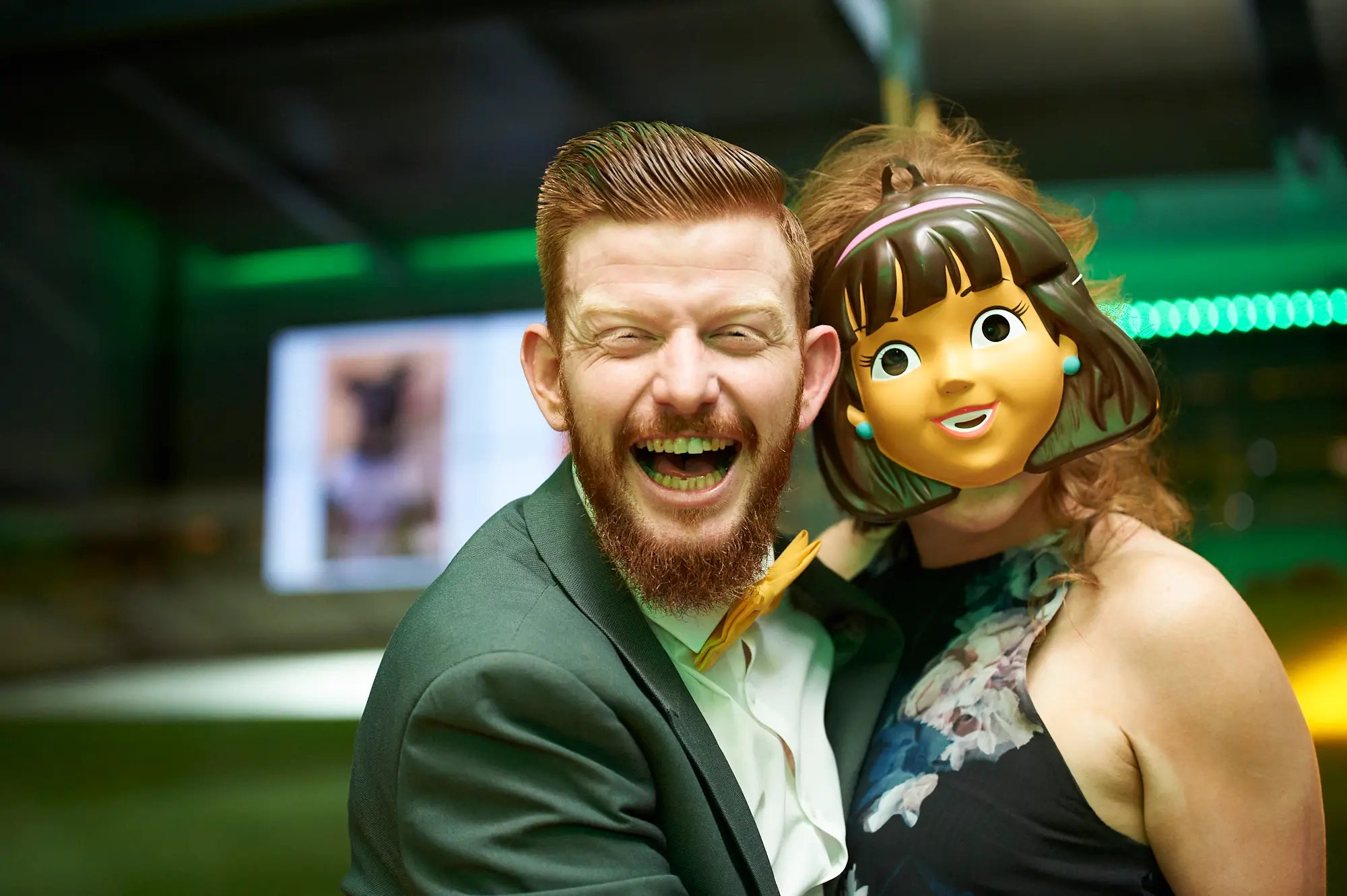 padraig pearses gac awards night mcg playful mask celebration A man with a red beard in a dark suit and yellow bow tie laughs ecstatically while posing next to a guest wearing a large, comical plastic mask of a cheerful female character. Both stand closely together, exuding playful energy. The modern MCG function room features vibrant green ambient lighting and sleek interiors. Guests in formal attire are enjoying a lively, celebratory moment, with the mask adding humor to the festive atmosphere, capturing the spontaneity, camaraderie, and joyful spirit of the gala event.