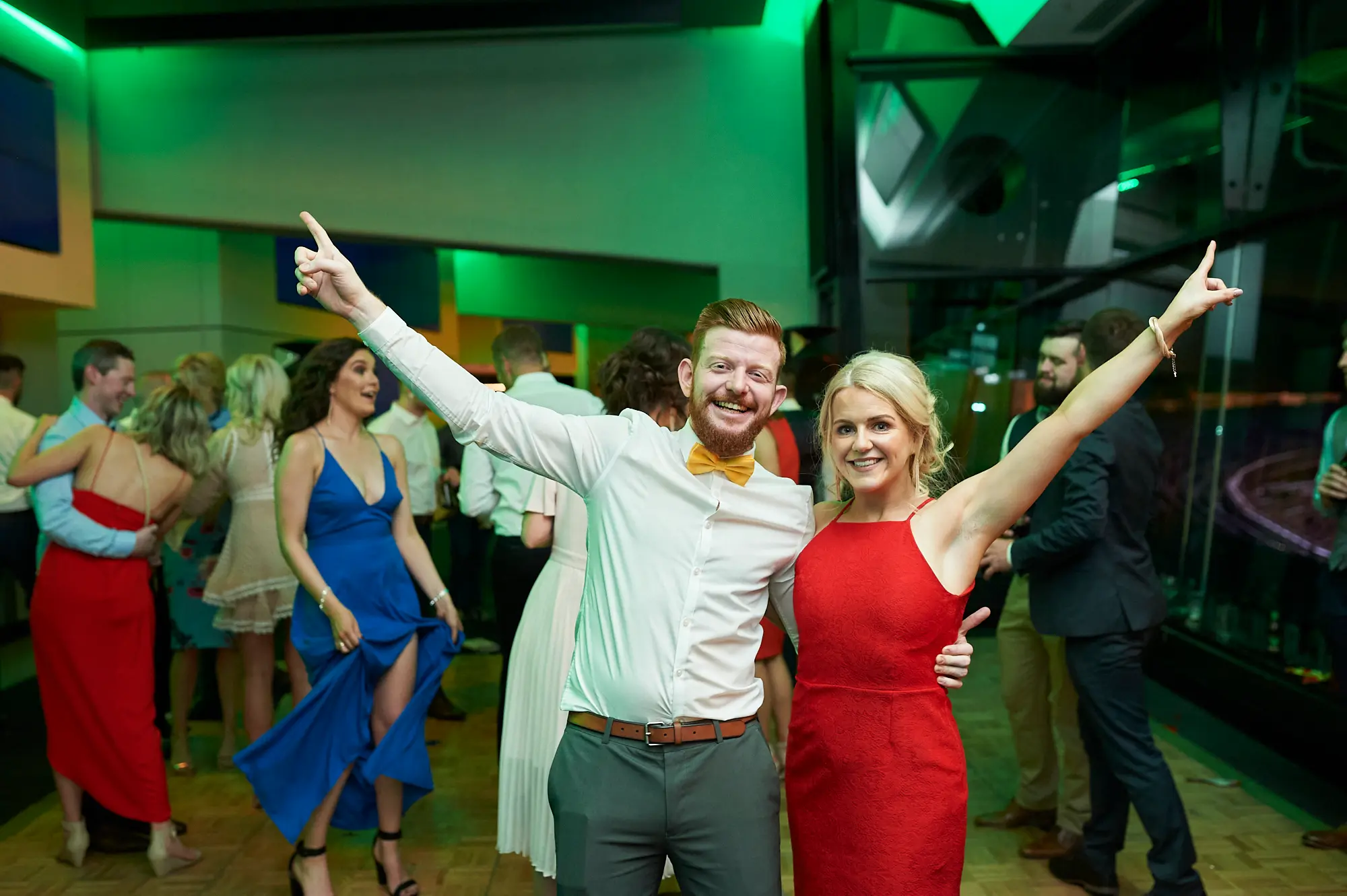 padraig pearses gac awards night mcg red dress celebration A man with a red beard in a white shirt and yellow bow tie and a woman in a vibrant red dress raise their arms enthusiastically during the Pearses Awards Night at the MCG, smiling broadly and capturing a lively moment of celebration. In the background, guests in formal attire mingle and dance, including a woman in a blue high-slit dress. The modern function room features floor-to-ceiling glass partitions with dark stadium seating beyond and vibrant green ambient lighting, creating a dynamic and festive atmosphere full of joy, camaraderie, and spirited enjoyment at the gala event.