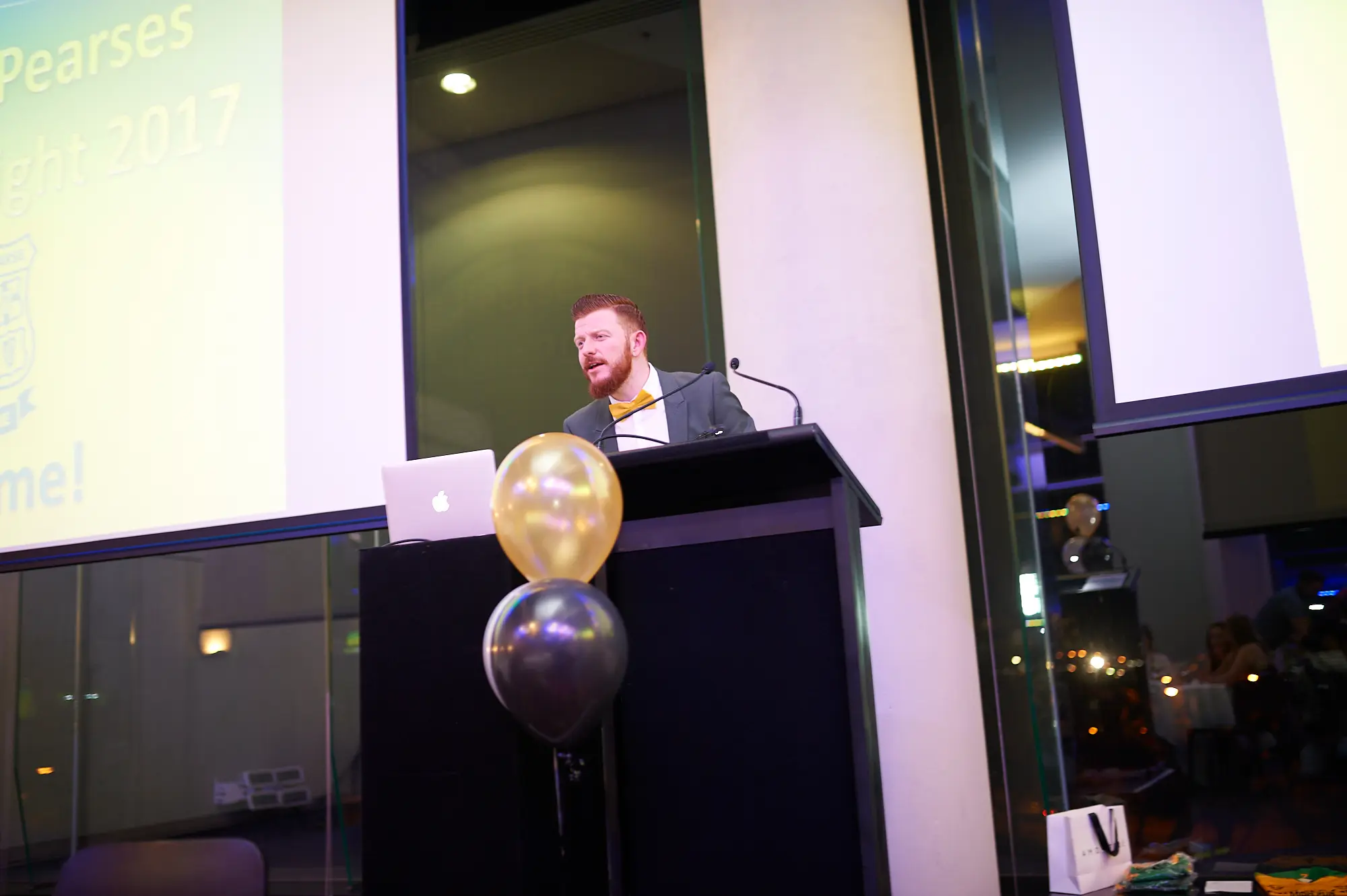 padraig pearses gac awards night mcg speaker lectern presentation A red-bearded man in a dark suit and yellow bow tie speaks animatedly at a black lectern during the Padraig Pearses GAC Awards Night at the MCG. Gold and gray balloons and an Apple laptop sit on the lectern, while a large projection screen displays event branding. Glass partitions reveal formally dressed guests seated at tables in the dimly lit background, creating a professional yet lively atmosphere as the speaker engages the audience with dynamic gestures and expressions.
