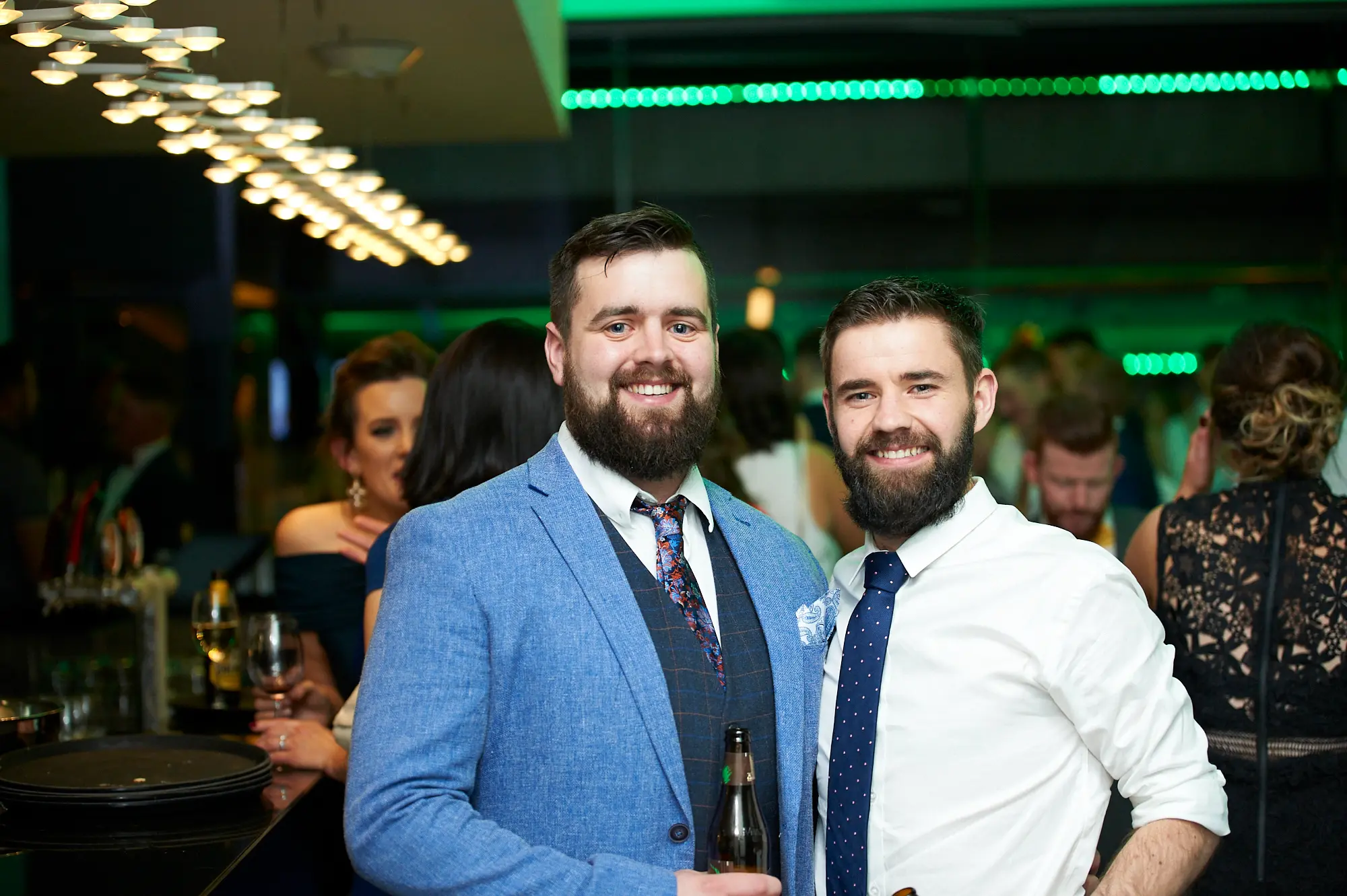 padraig pearses gac awards night mcg two men bar Two men in formal attire pose together at the Pearses Awards Night at the MCG, smiling warmly at the camera. One holds a beer bottle while the other stands casually. Behind them, guests mingle near a modern bar area illuminated with vibrant green ambient lighting. A woman in an off-shoulder dark blue dress holds a wine glass, adding to the social atmosphere. Dark plates on the bar and sleek contemporary décor highlight the sophisticated event setting, capturing a moment of relaxed camaraderie, elegance, and festive celebration among the attendees.