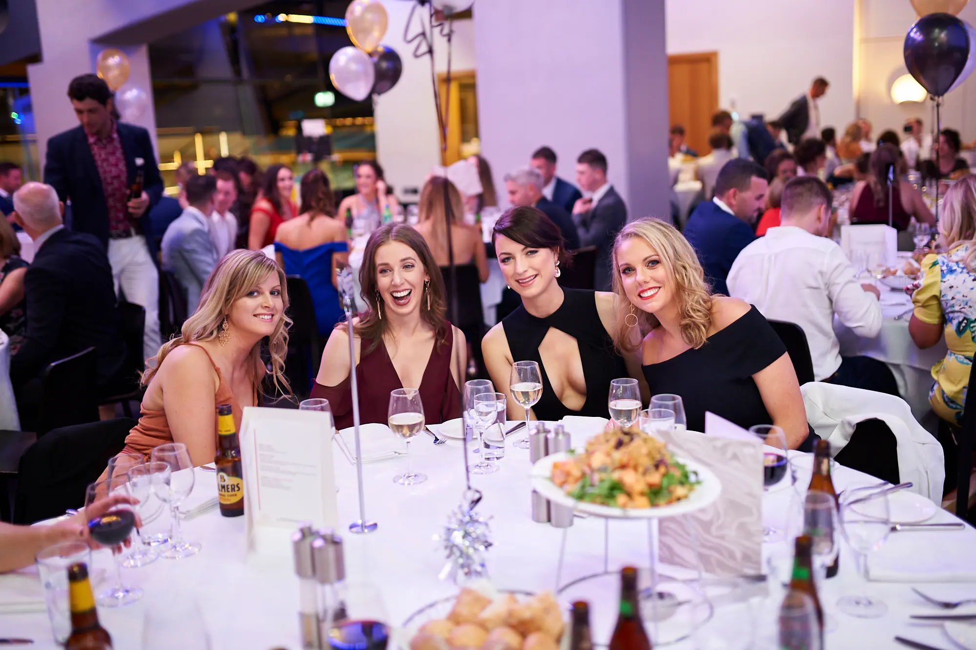 padraig pearses gac awards night mcg women dinner table laughter Four women share a joyful moment at a dinner table during the Padraig Pearses GAC Awards Night & Gala Dinner at the MCG. One woman laughs heartily while the others smile warmly, holding wine glasses or with drinks nearby. The large event hall features round tables with white tablecloths, black, gold, and white balloons, and warmly lit surroundings. Other formally dressed guests mingle and dine in the background. The scene captures a festive, celebratory, and social atmosphere, highlighting friendship, happiness, and shared enjoyment during the gala dinner.
