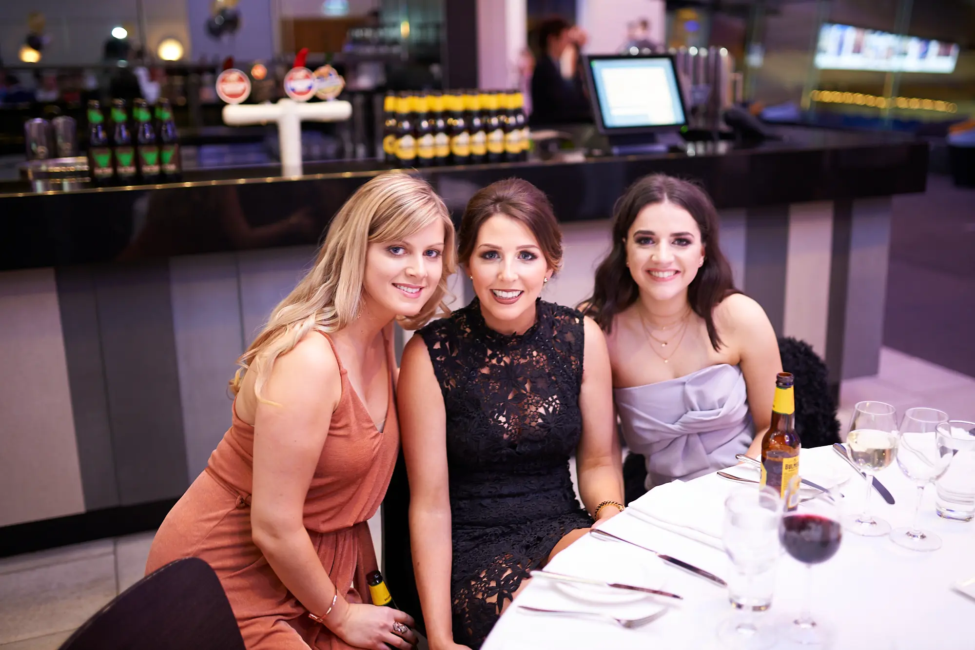 padraig pearses gac awards night mcg women dinner table peach black grey Three women pose and smile warmly at a dinner table during the Padraig Pearses GAC Awards Night & Gala Dinner at the MCG. The woman on the left wears a peach-colored dress, the center woman wears a black lace dress, and the woman on the right wears a light grey strapless dress. The table features wine glasses, cutlery, and a small beer bottle. A modern bar with a sleek black counter and striped panels is visible in the background. The scene captures a festive, elegant, and social atmosphere as guests enjoy drinks, laughter, and camaraderie during the gala dinner.