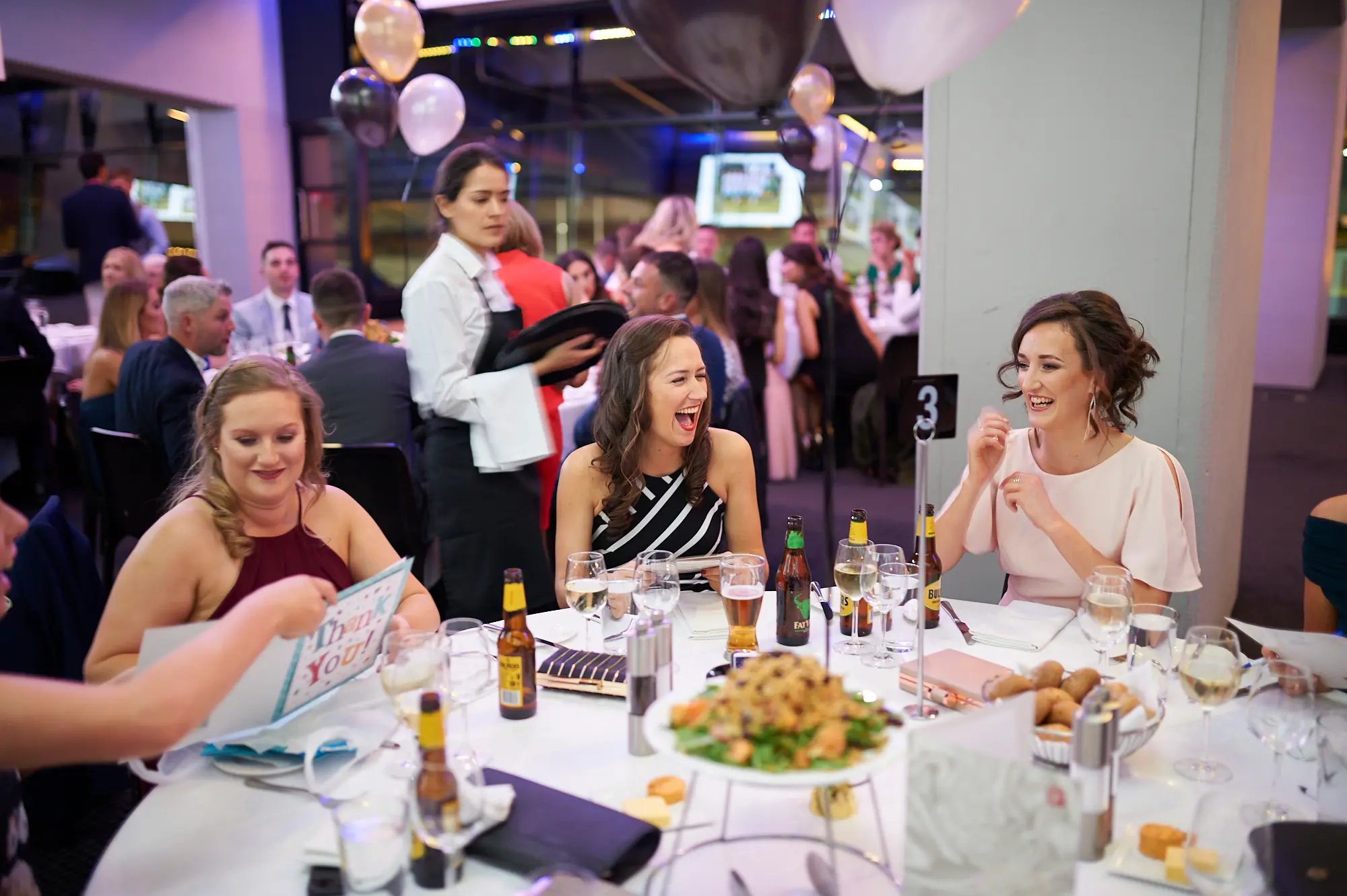 padraig pearses gac awards night mcg women dinner table striped pink maroon Three women share a lively moment at a dinner table during the Padraig Pearses GAC Awards Night & Gala Dinner at the MCG. The center woman in a black and white striped dress laughs heartily, while the woman in a light pink dress smiles broadly, and the woman in a maroon dress looks down with a pleasant expression. The table is set with wine glasses, cutlery, and beer bottles. White tablecloths, decorative gold, black, and white balloons, and warmly lit surroundings create a festive, social, and celebratory atmosphere as guests enjoy dinner, drinks, and camaraderie.