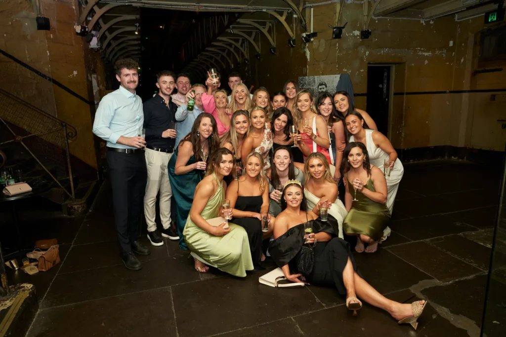 svg%3E A large group of guests pose together for a joyful photograph during the Padraig Pearses GAC Awards Night and End of Year Celebration at Melbourne Old Gaol. Dressed in formal evening wear and holding drinks, the group gathers casually across the floor and steps, laughing and smiling. Framed by the gaol’s rugged bluestone walls, archways, and moody lighting, the image captures the lively, social spirit of this community-focused celebration in a historic Melbourne venue.