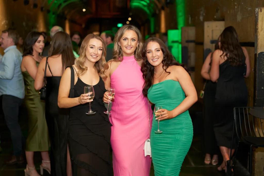 svg%3E Three women stand close together, smiling at the camera while holding wine glasses during the Padraig Pearses GAC Awards Night and End of Year Celebration at Melbourne Old Gaol. Framed by the venue’s historic, industrial architecture and soft green uplighting, the scene captures a relaxed and social moment. Other guests mingle in the background in elegant evening attire, reflecting the warm, celebratory atmosphere of this community-focused event.
