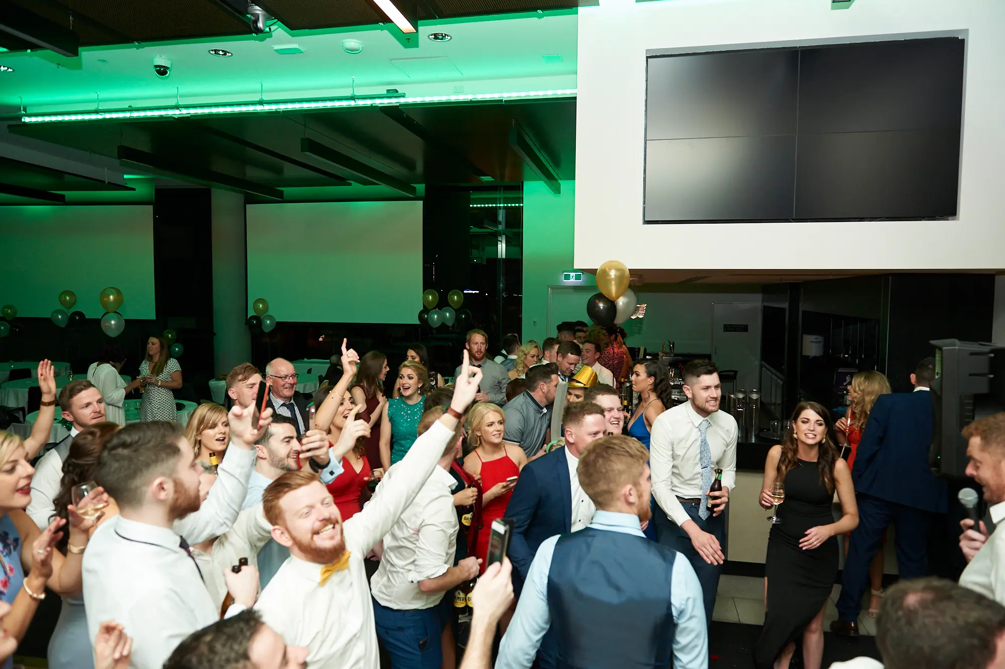 pearses awards night mcg dance floor celebration wide shot A bearded man in a white shirt and tie laughs ecstatically, holding a beer bottle, while a woman in a black dress cheers with a phone raised. Guests in suits and evening gowns surround them, dancing and celebrating. Vibrant green ambient lighting, glass partitions, and stadium seating in the background highlight the energetic and joyful party atmosphere at the MCG gala.