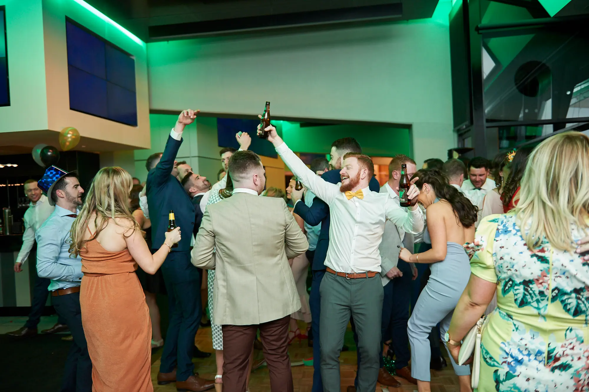 pearses awards night mcg dance floor celebration Guests celebrate energetically on the dance floor during the Pearses Awards Night at the MCG, with arms raised, drinks in hand, and wide smiles. A man in a white shirt and yellow bow tie laughs ecstatically while holding a beer bottle, while others in formal attire cheer and dance around him. Women in vibrant dresses join the revelry, and a man in a blue suit raises his arm in excitement. The modern venue features vibrant green ambient lighting, glass partitions, and blue display screens, capturing the dynamic energy, joyful interactions, and spirited atmosphere of the gala event.