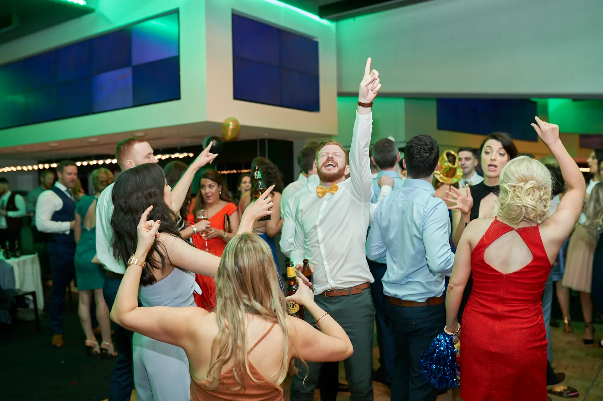 pearses awards night mcg dance floor energy celebration Guests celebrate energetically on the dance floor during the Pearses Awards Night at the MCG, with laughter, raised arms, and drinks in hand. A bearded man in a white shirt and yellow bow tie points upwards while holding a beer bottle, capturing a spontaneous moment of joy. Nearby, a woman in a black dress cheers with her phone, while others hold drinks or decorative items. The modern venue features tiered stadium seating, green ambient lighting, mirrored partitions, and large blue screens, creating a dynamic and festive atmosphere filled with social interaction, fun, and excitement.