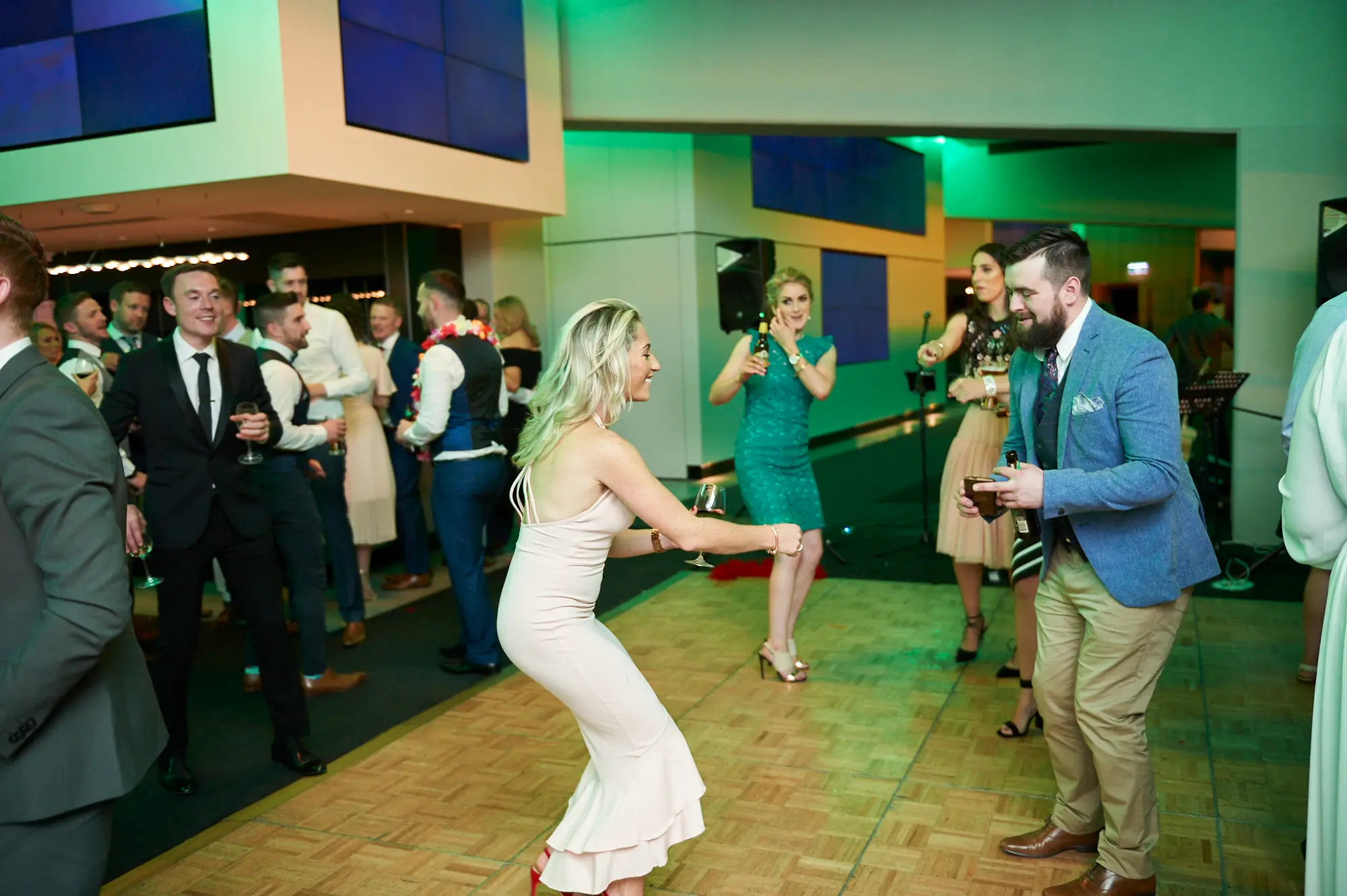 pearses awards night mcg dance floor energy Guests enjoy lively dancing during the Pearses Awards Night at the MCG, with a woman in a light pink dress leaning back energetically while smiling at a man in a blue suit holding drinks. A woman in a vibrant green dress dances nearby, and other formally dressed attendees mingle and cheer across the wooden dance floor. The modern venue glows with vibrant green ambient lighting, with blue display screens adding a contemporary touch. The scene captures the excitement, camaraderie, and spirited energy of a formal gala that has evolved into a joyful, interactive celebration on the dance floor.