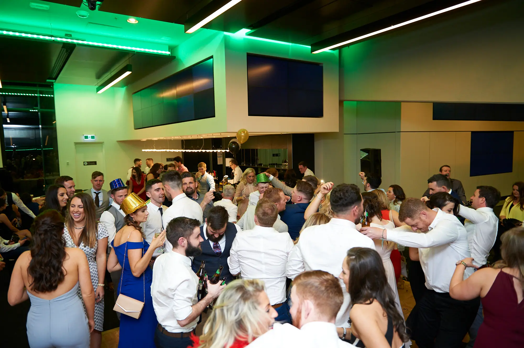pearses awards night mcg dance floor party celebration Guests at the Pearses Awards Night at the MCG celebrate energetically on the dance floor. Men in suits and novelty party hats hold beer bottles and laugh, while women in vibrant evening dresses smile, cheer, and raise their hands. The modern venue features large glass partitions, tiered stadium seating visible in the background, and dynamic green ambient lighting. The scene captures the peak of the gala’s party atmosphere, with guests mingling, dancing, and enjoying drinks, highlighting the joyous, lively, and social energy of the evening.