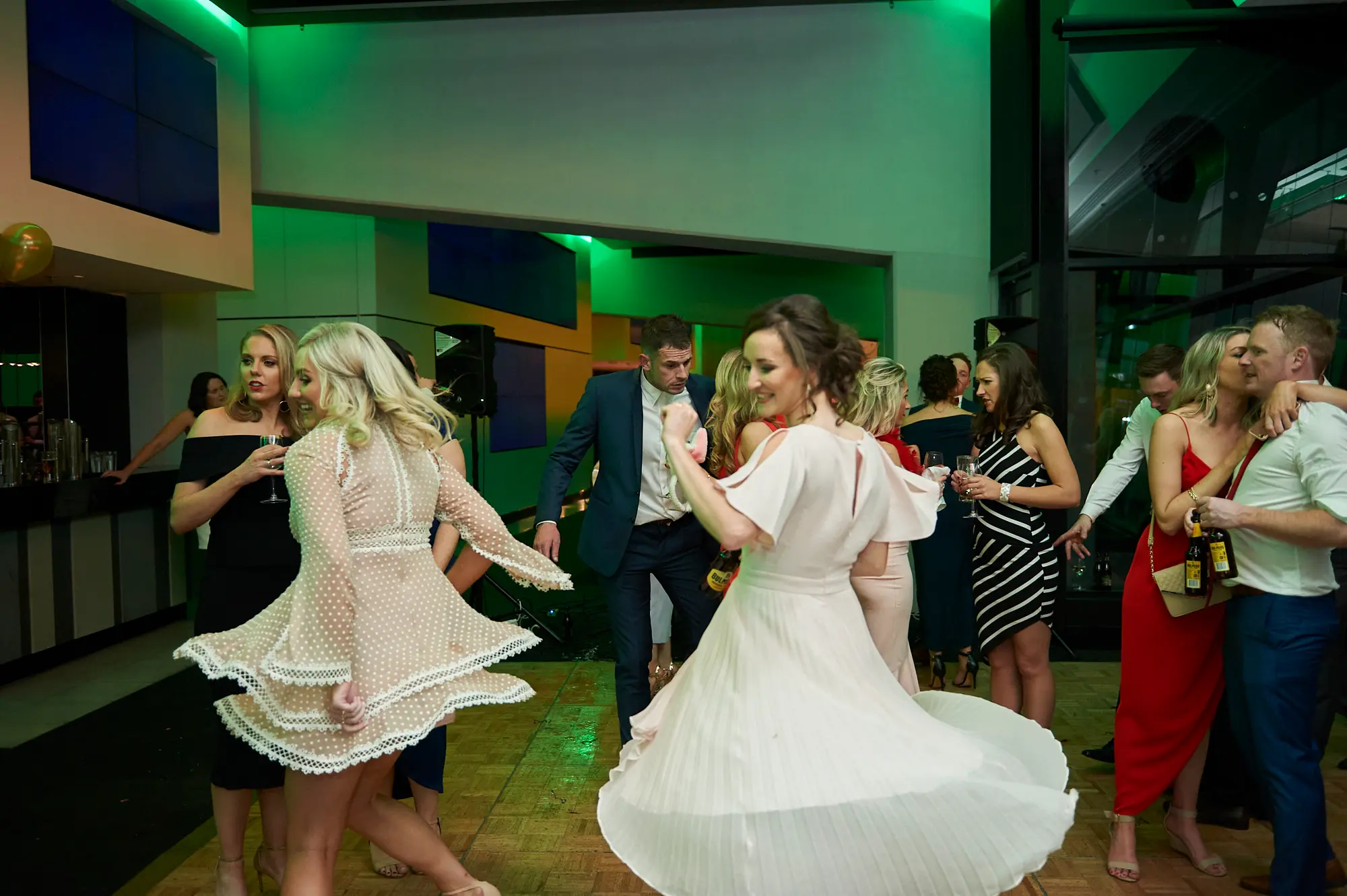 pearses awards night mcg dance floor spin dresses Two women are captured mid-spin on the dance floor, their elegant dresses fanning out dramatically as they smile and hold drinks. Around them, guests in formal attire mingle, dance, and cheer, creating a lively and joyful atmosphere. Vibrant green ambient lighting, modern glass partitions, and the MCG stadium seating in the background enhance the dynamic, celebratory mood of the gala event.
