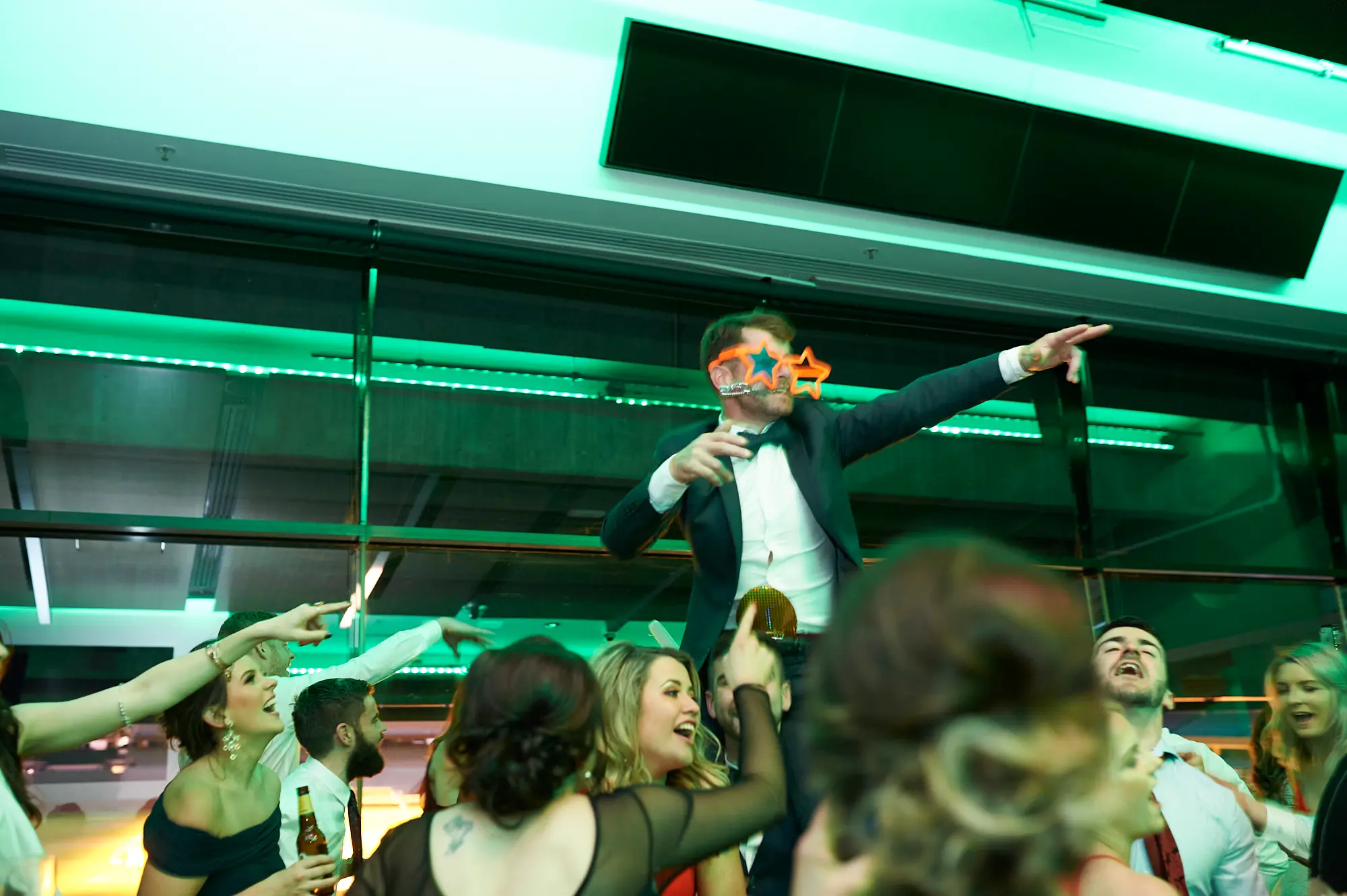 pearses awards night mcg dance floor star glasses Guests erupt in laughter and cheers as a man wearing orange star-shaped novelty glasses is lifted above the dance floor, arms outstretched in celebration. Around him, men and women in formal attire point, smile, and hold drinks, creating a scene full of energy, spontaneity, and joy. The modern function room, green ambient lighting, and MCG stadium seating in the background enhance the dynamic, party atmosphere of the gala event.