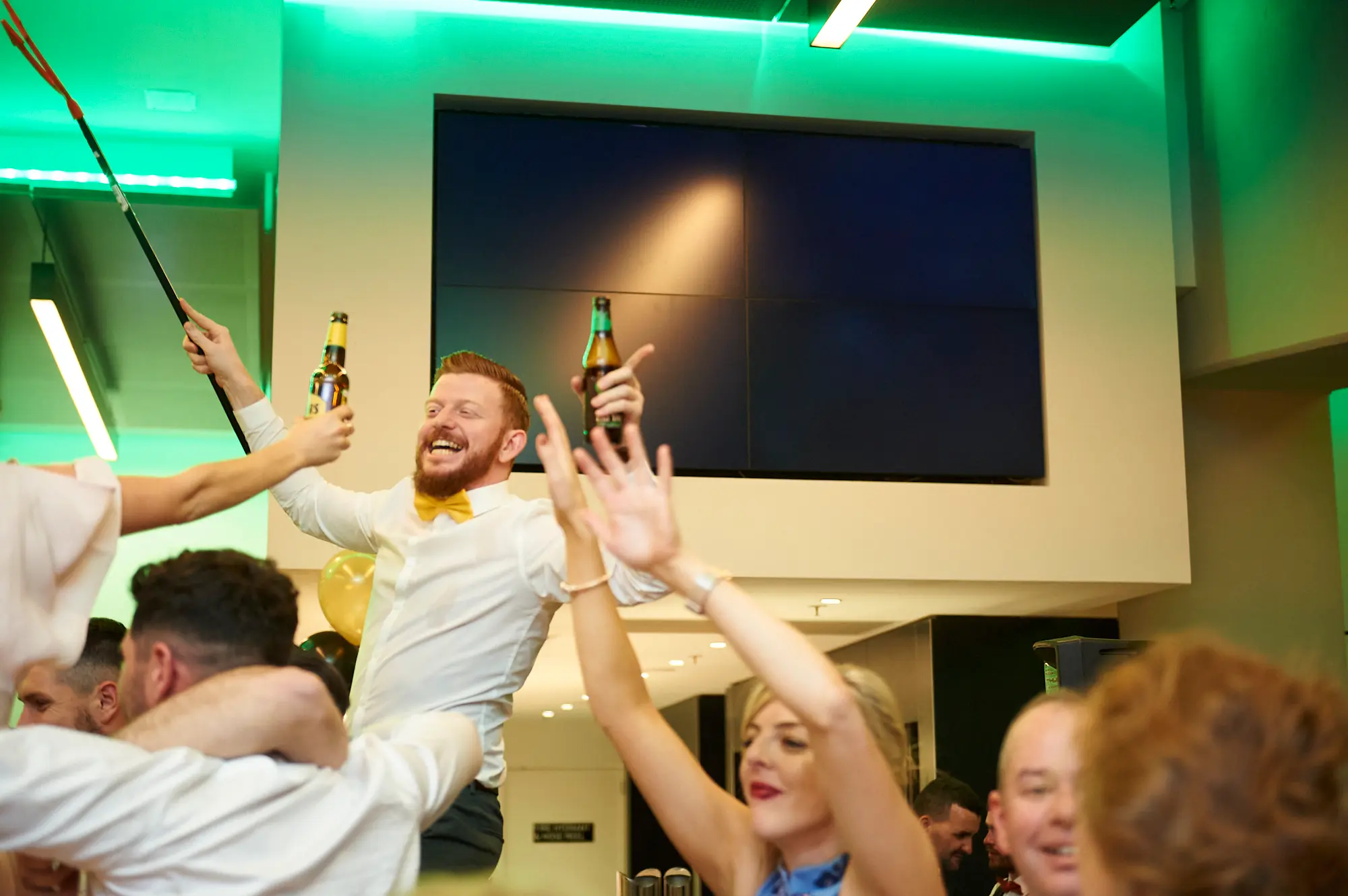 pearses awards night mcg dance floor toast A bearded man in a white shirt and yellow bow tie raises a beer bottle with wide-eyed excitement while another person clinks bottles with him. A woman in a blue dress cheers nearby, and guests in suits and evening gowns mingle and dance. Vibrant green lighting and modern venue elements at the MCG highlight the lively, joyful celebration on the dance floor.