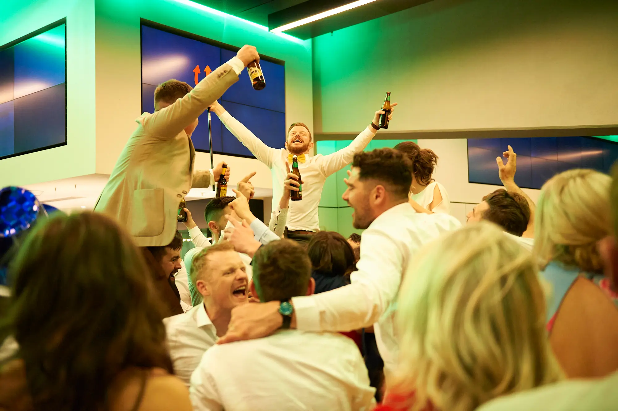 pearses awards night mcg dance floor triumph A bearded man in a white shirt and yellow bow tie stands on an elevated surface with arms raised, holding two beer bottles, laughing broadly. Guests around him cheer, smile, and raise their hands in excitement. Women in colorful dresses and men in suits mingle and dance under vibrant green lighting at the MCG, capturing a moment of peak celebration and joy on the dance floor.