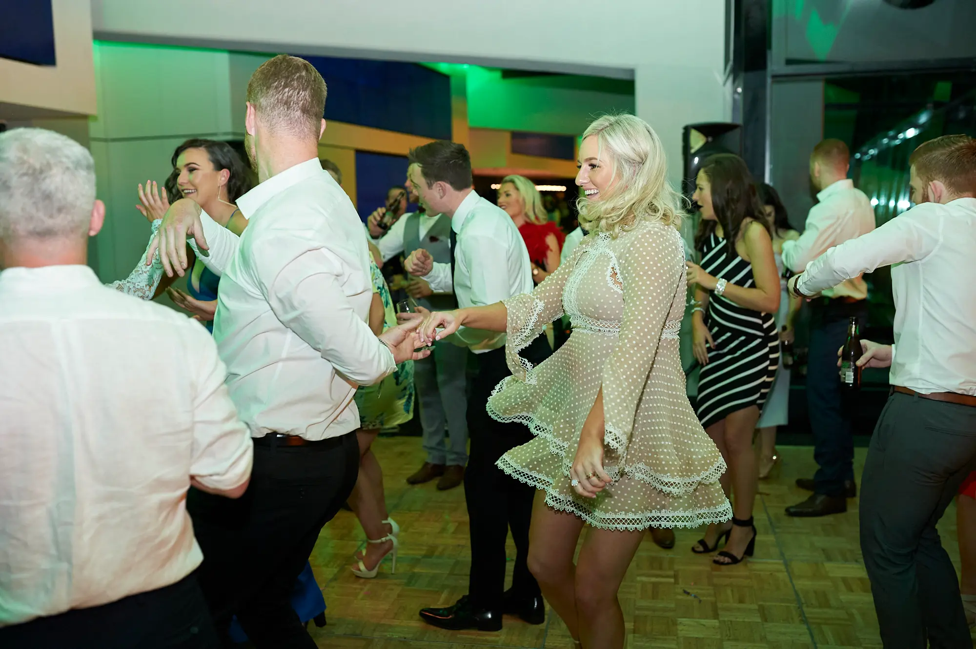 pearses awards night mcg dance floor twirl A woman in a beige polka-dot dress is mid-twirl on the dance floor, holding hands with a man in a white shirt, smiling broadly. Around them, guests in formal attire mingle, dance, and cheer, holding drinks. Vibrant green ambient lighting, modern glass partitions, and the MCG stadium seating in the background enhance the lively, joyful, and celebratory atmosphere of the gala event.